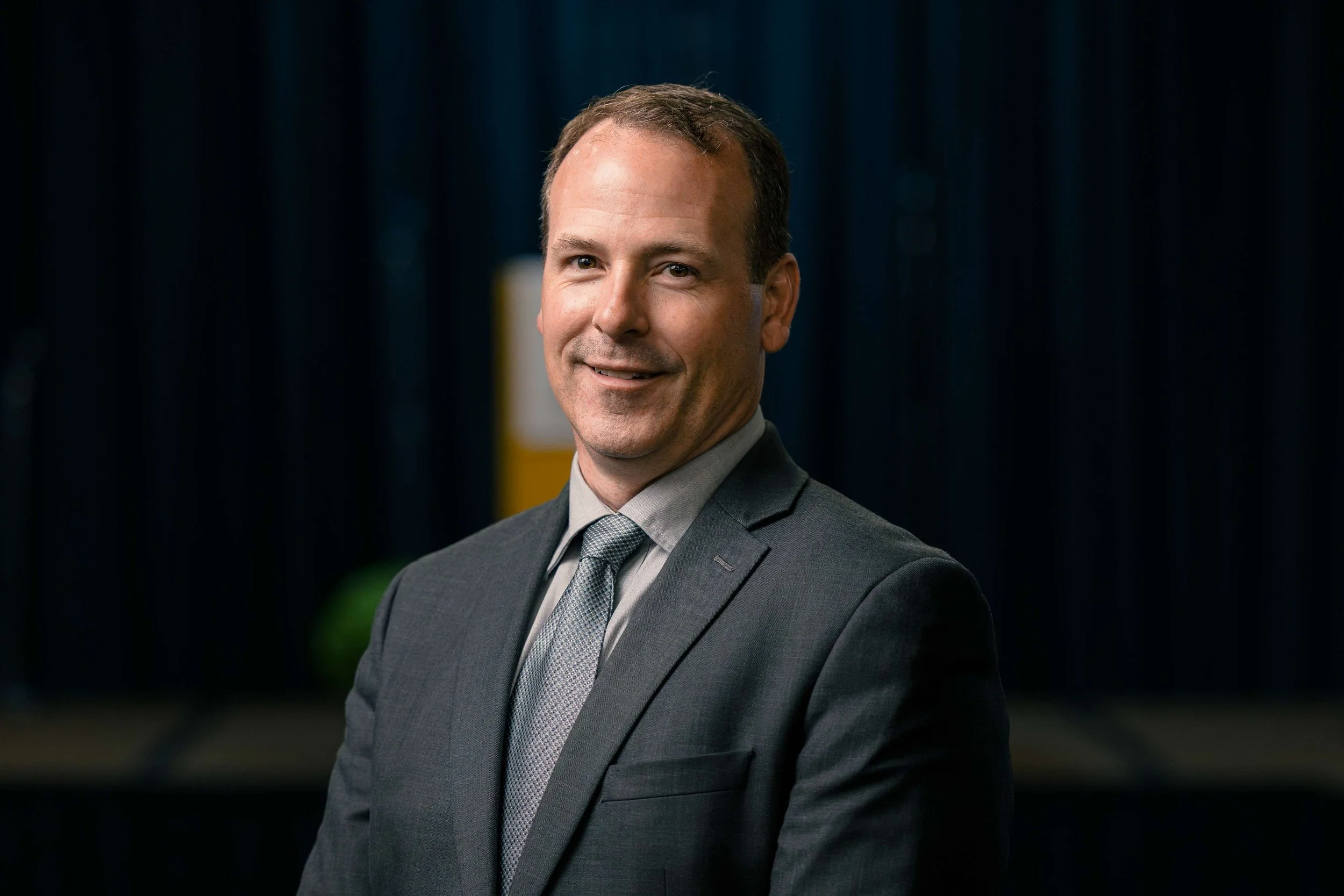 A man in a gray suit and tie smiling at the camera, standing indoors with dark curtains and a blurred background.
