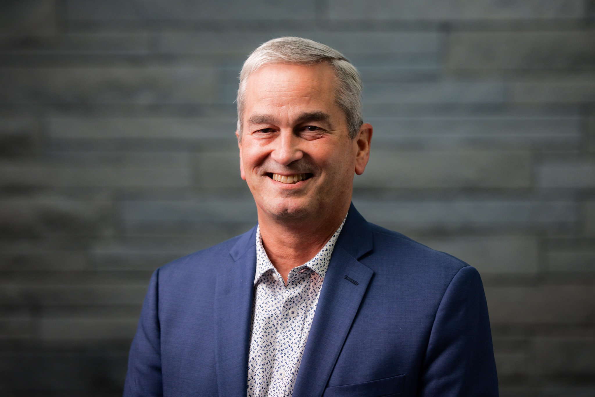 Headshot of a middle-aged man with gray hair, smiling, wearing a blue suit jacket and a white patterned shirt, standing in front of a gray textured background.