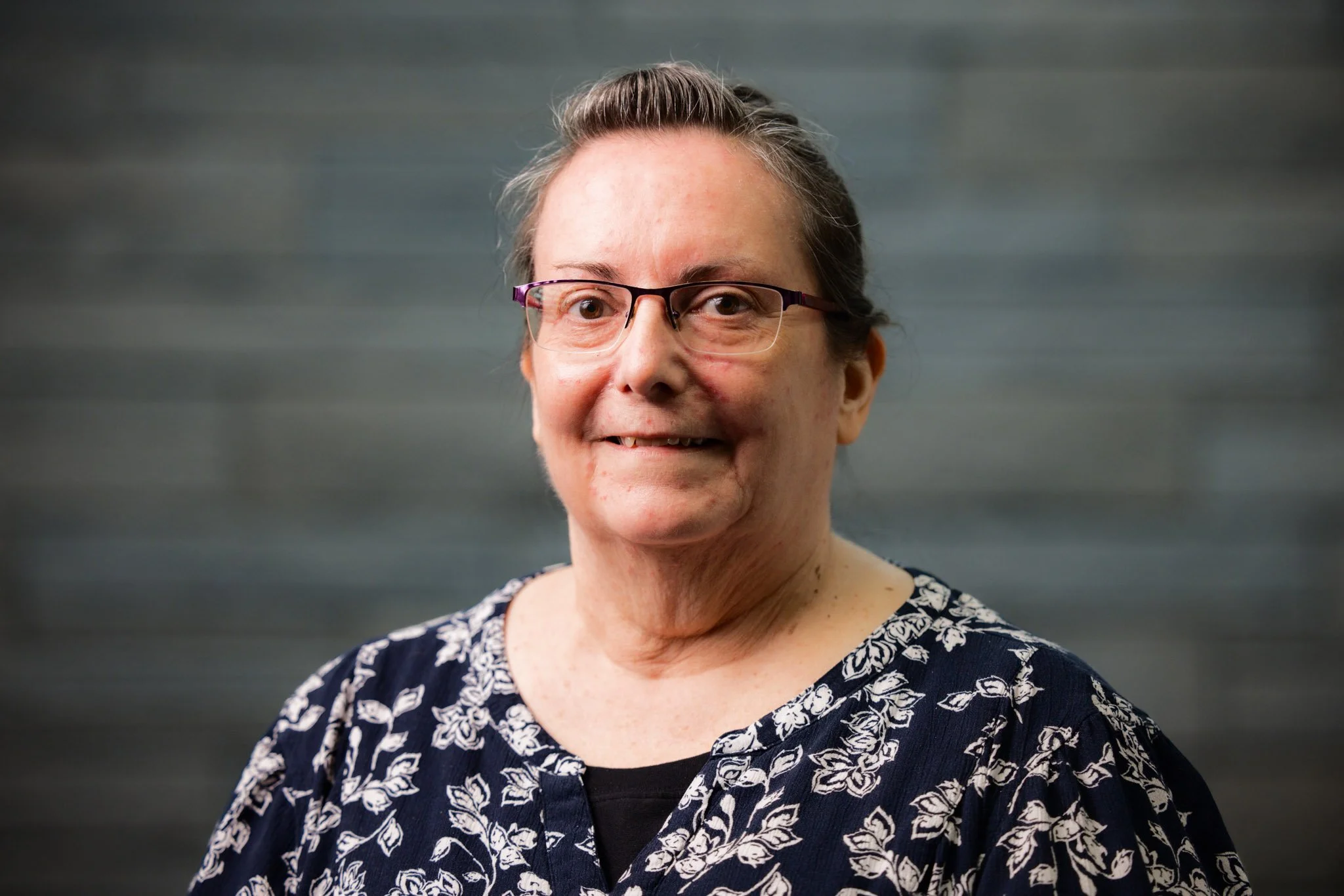 Portrait of an elderly woman with short gray hair, glasses, and a floral dark blue top, smiling against a blurred gray background.
