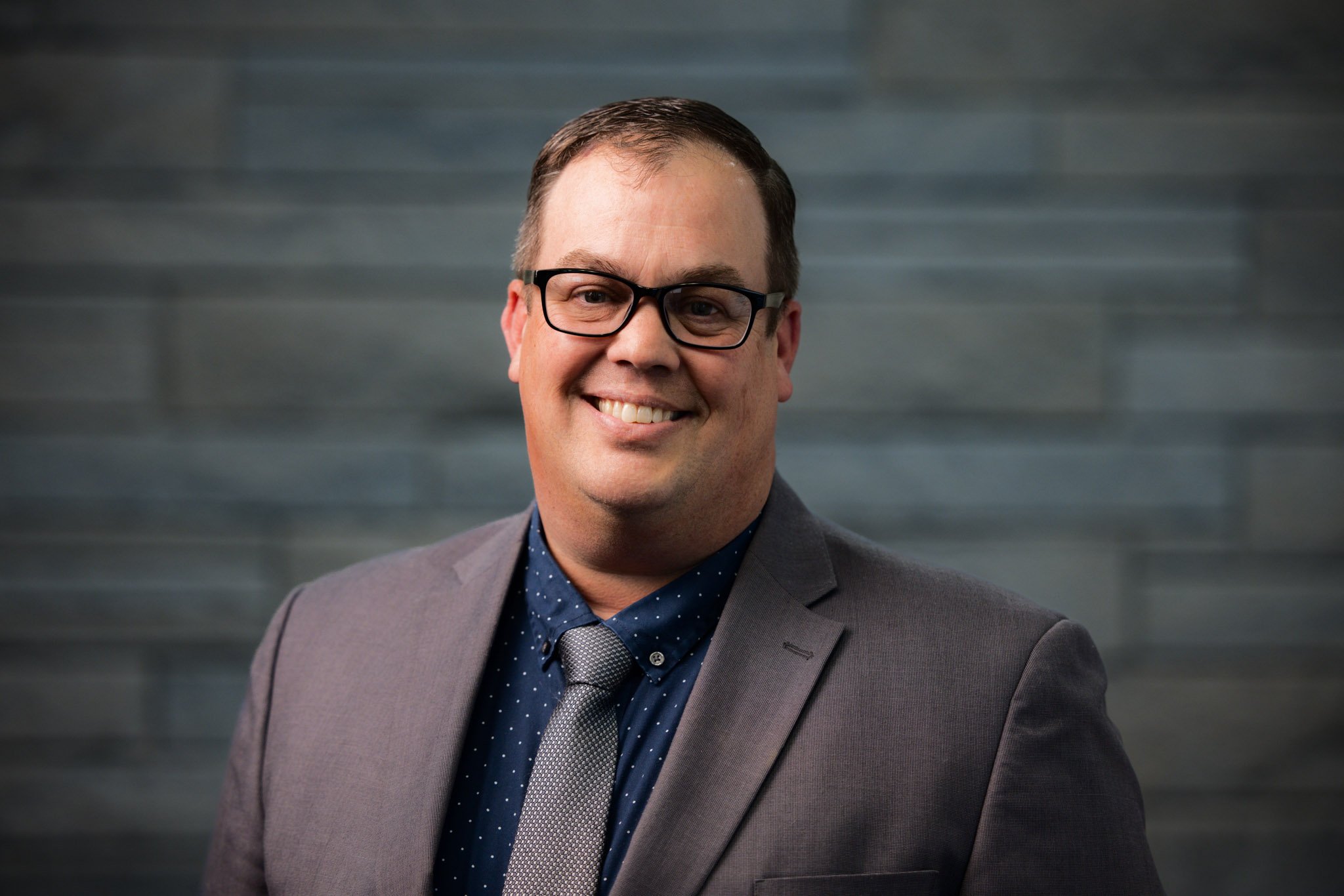A smiling man in a gray suit, navy blue shirt with white dots, and gray tie, wearing black glasses, standing against a dark gray textured wall background.