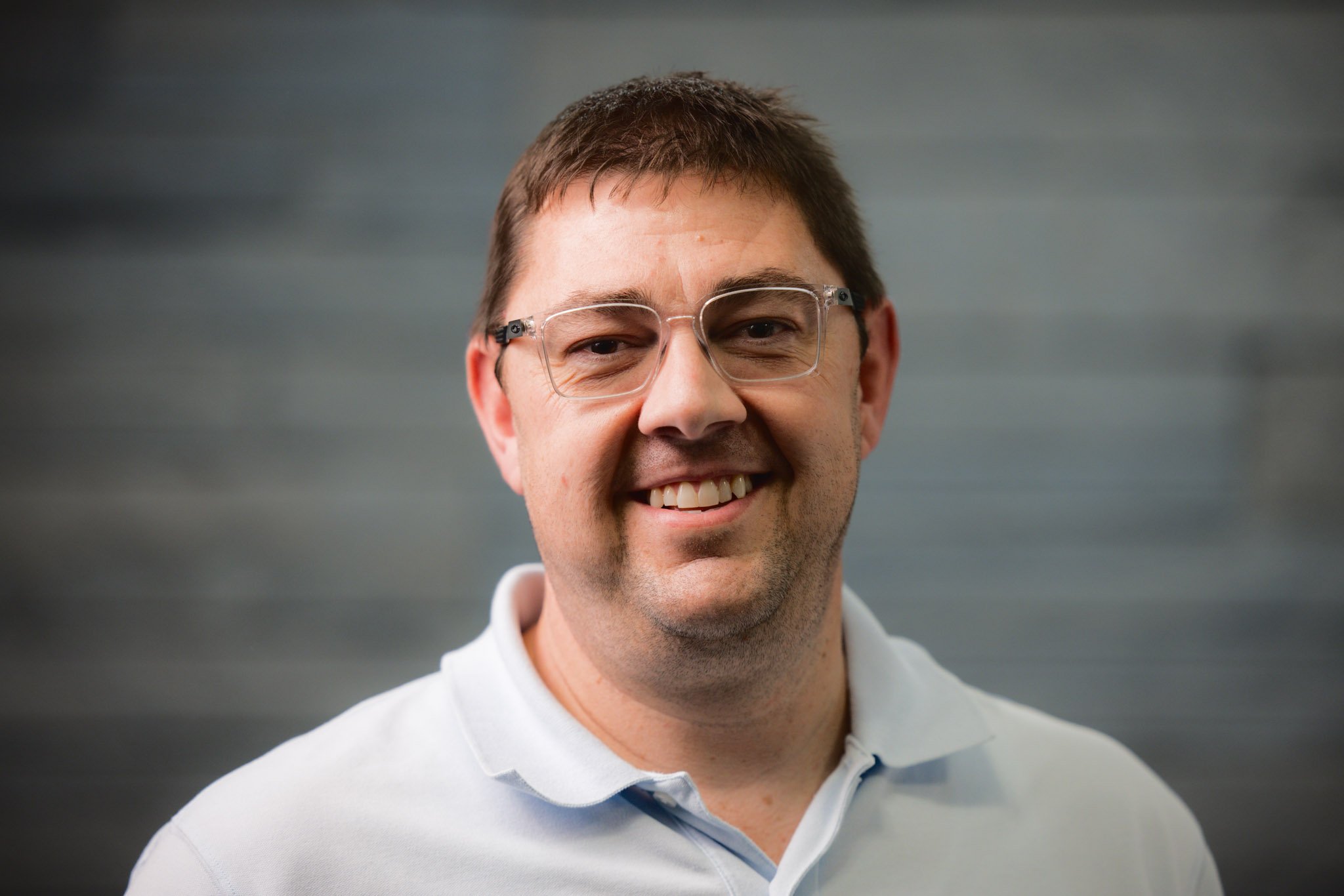 A smiling man with short brown hair, wearing glasses and a white polo shirt, standing in front of a blurred gray background.