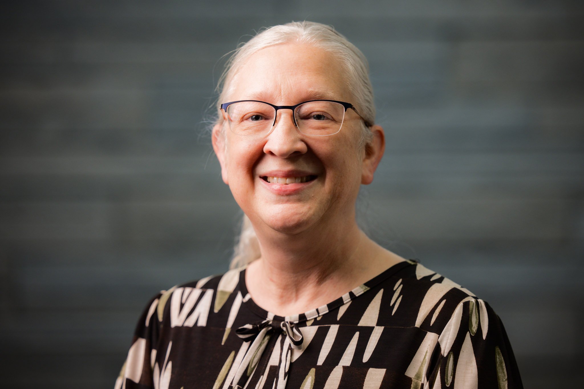 Portrait of an elderly woman with gray hair, glasses, and a patterned blouse, smiling against a dark background.