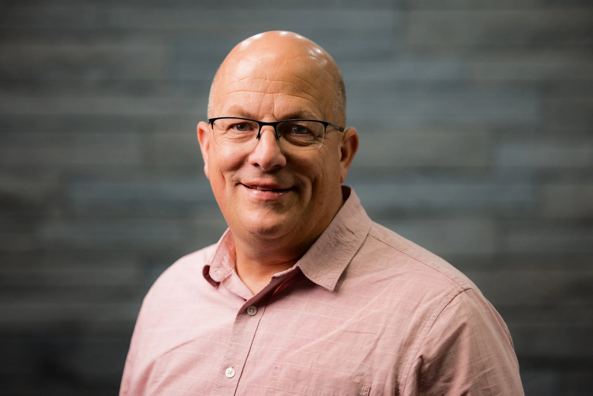 A middle-aged man with glasses and a bald head, wearing a light pink button-up shirt, smiling, with a gray blurred background.