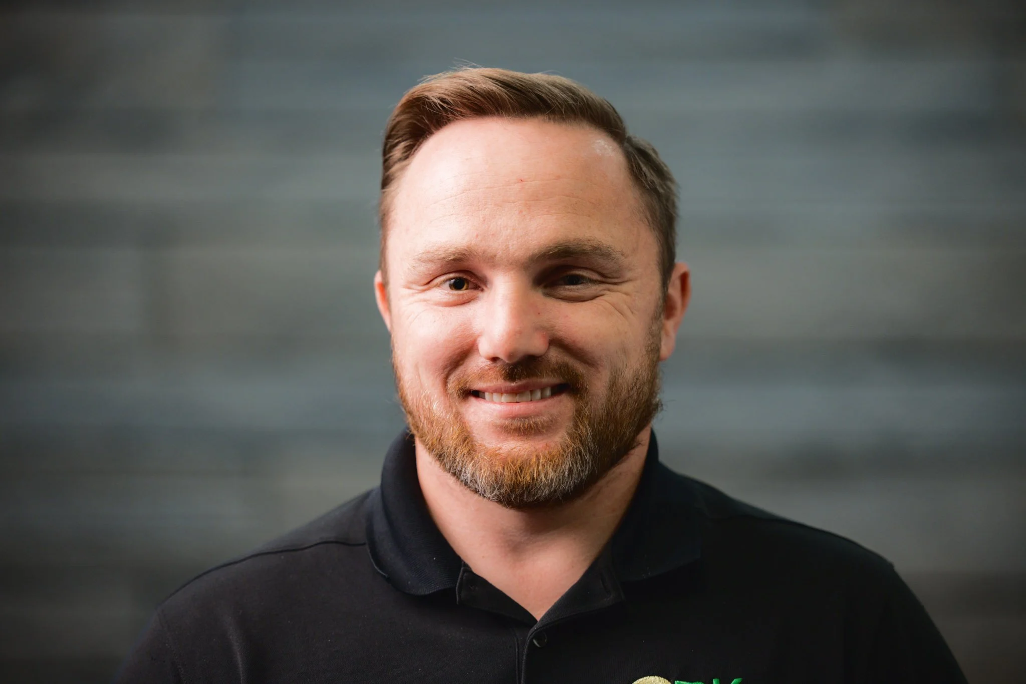 A smiling man with light brown hair and a beard, wearing a black polo shirt with a logo, standing against a blurred gray background.