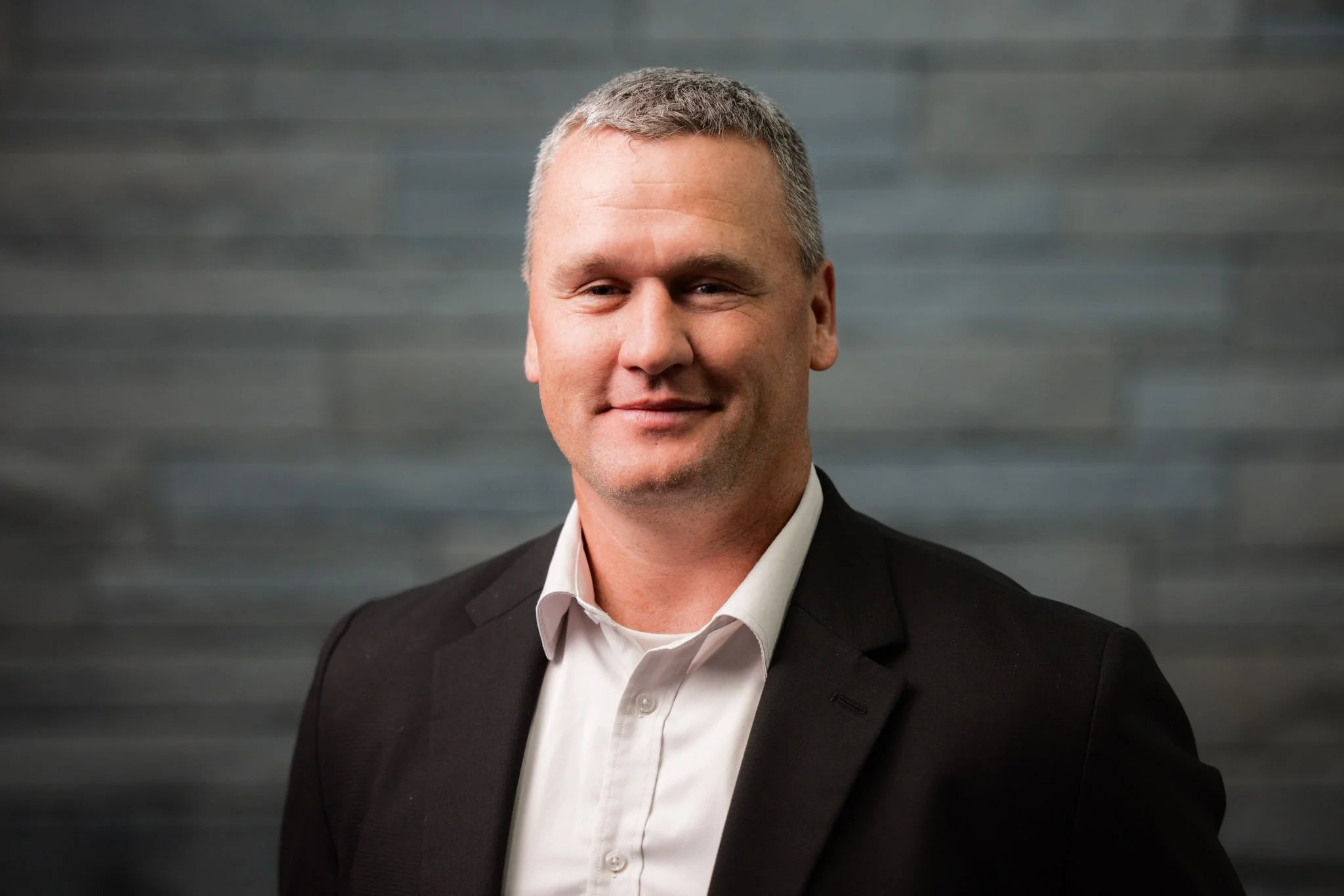 Portrait of a middle-aged man with short gray hair, wearing a black suit jacket and white dress shirt, smiling softly, against a gray textured background.