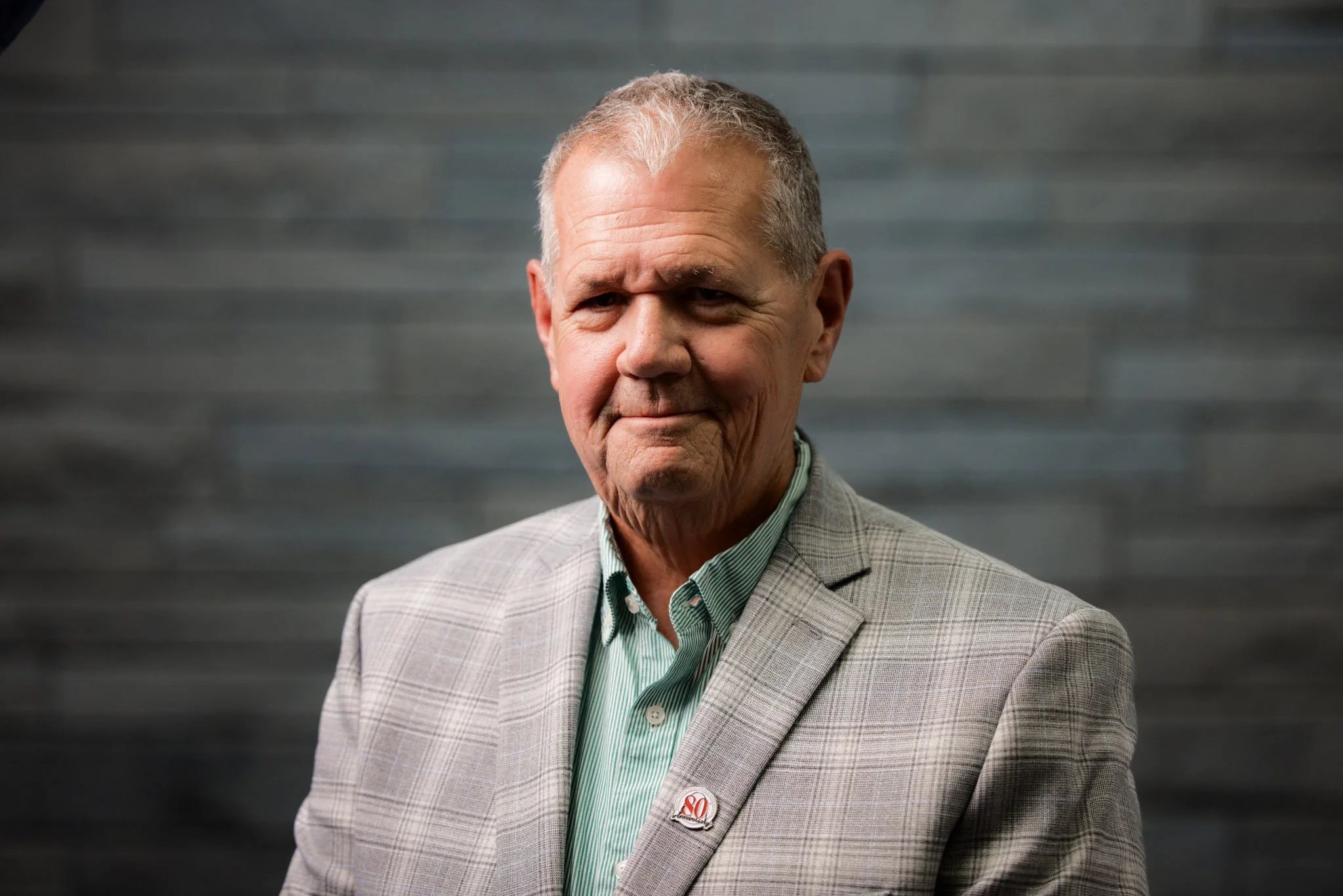 An elderly man with gray hair and a slightly wrinkled face wearing a light gray plaid blazer over a green striped button-up shirt, standing against a dark gray background.