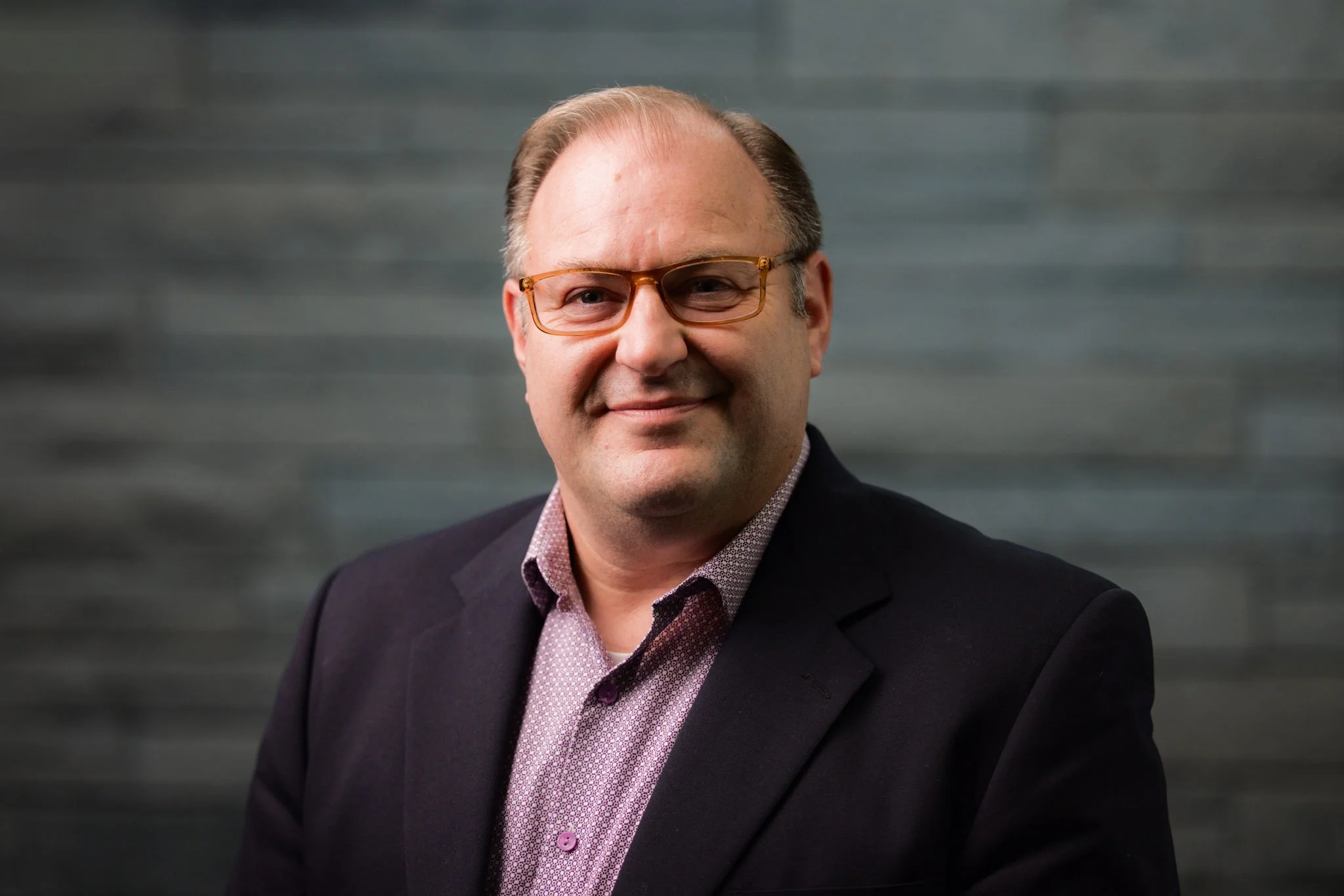 Headshot of a smiling man with glasses wearing a dark blazer and a patterned button-up shirt, standing against a gray, textured background.