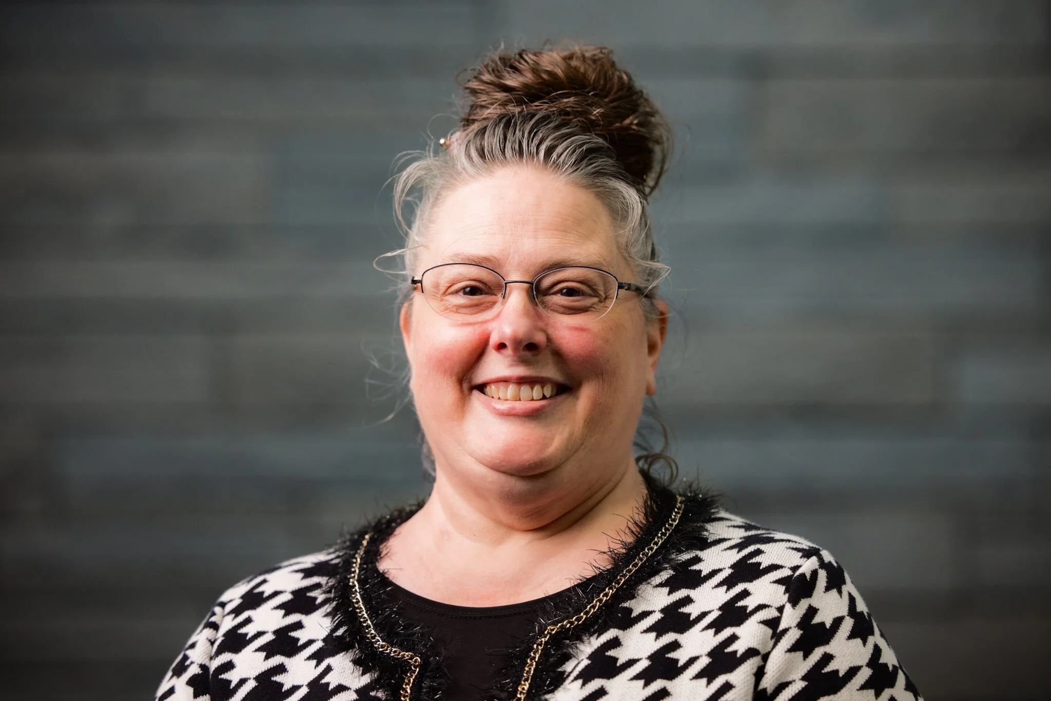 Smiling middle-aged woman with glasses and gray hair in a bun, wearing a black and white houndstooth patterned top, standing against a gray textured background.