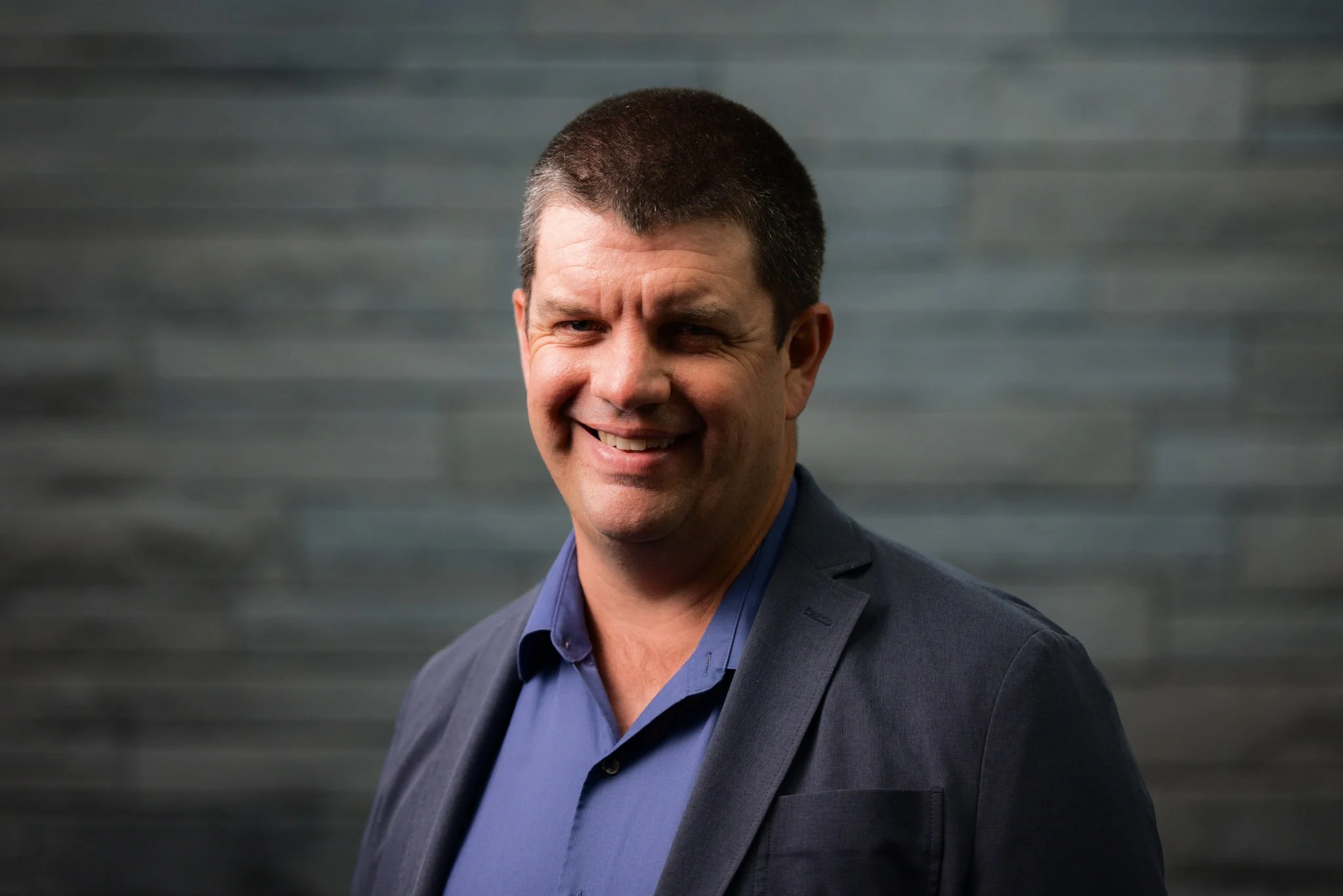 A man smiling, wearing a dark blazer and a blue shirt, standing in front of a dark gray brick wall.