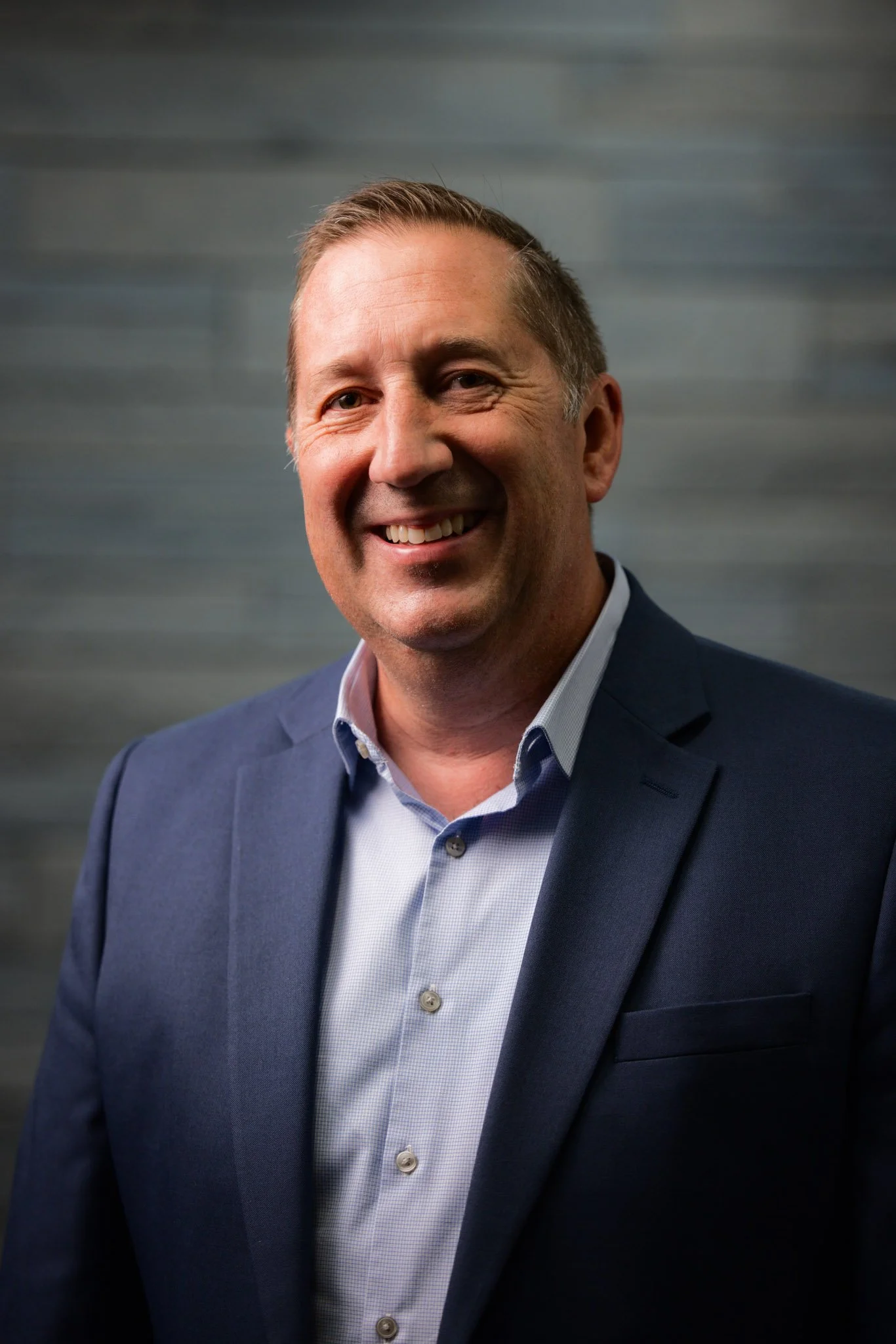 A man in a blue suit smiling for a portrait against a dark gray textured background.