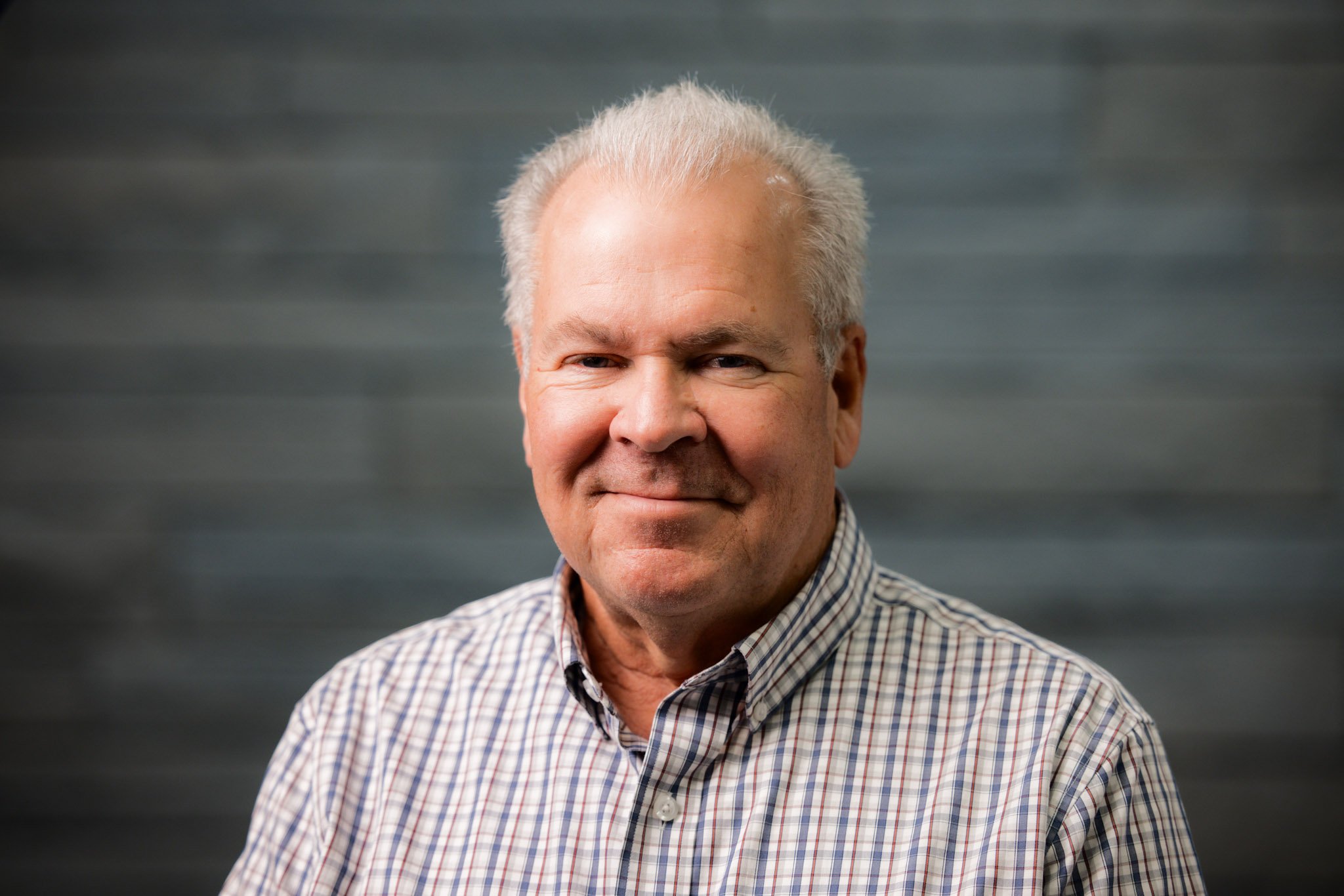 Portrait of an older man with gray hair, wearing a checkered shirt, smiling against a dark, blurred background.