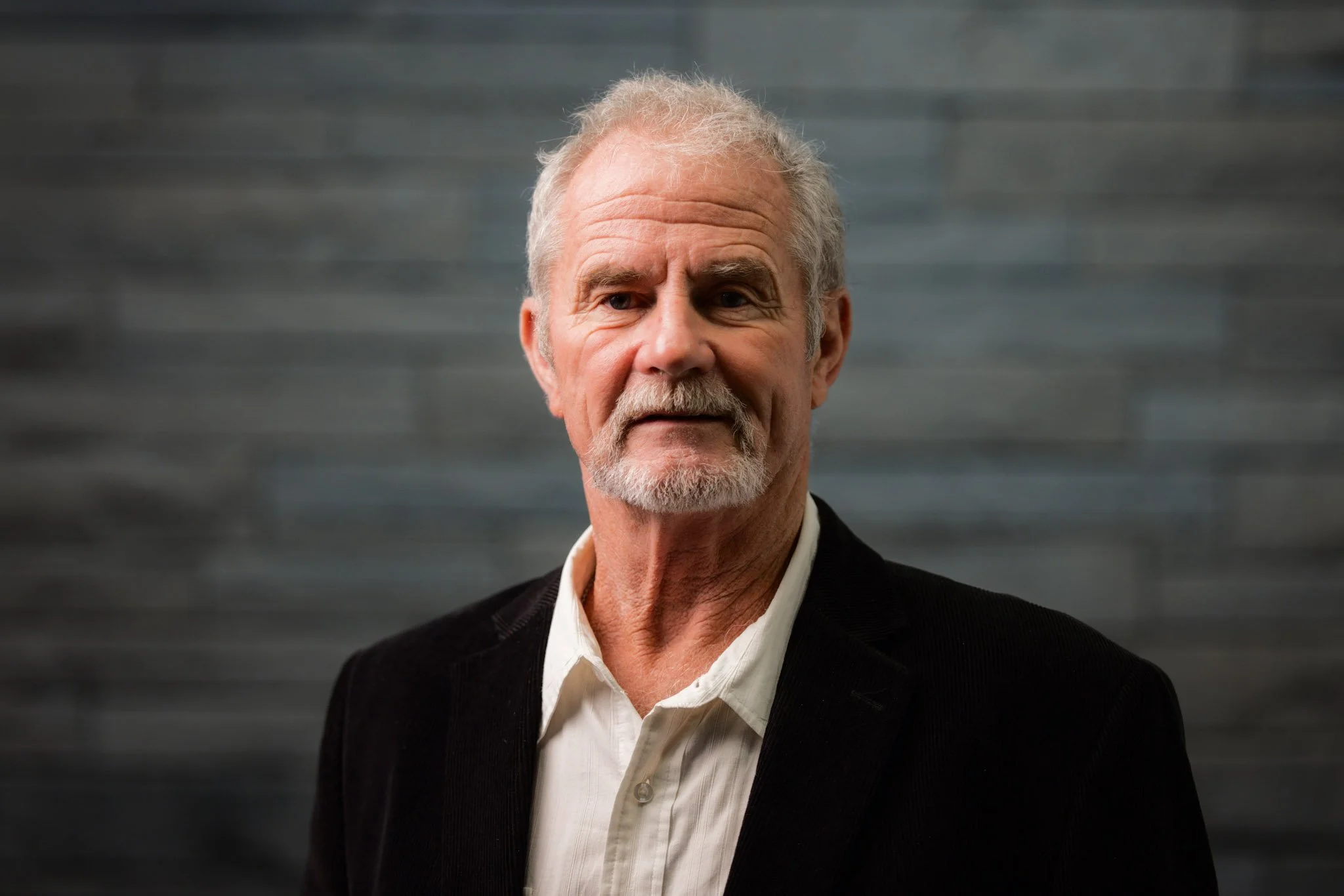 An older man with gray hair and a beard, wearing a white shirt and black blazer, standing against a dark gray background.