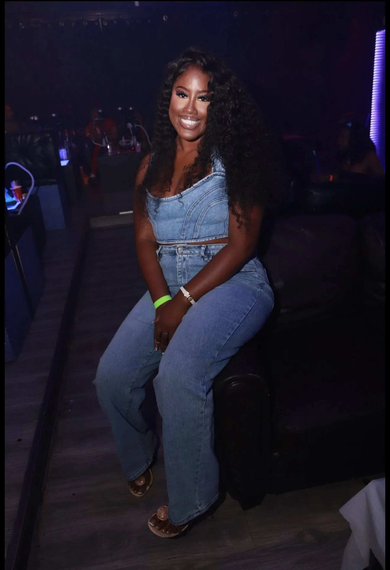 A woman sitting on a black couch in a dimly lit venue, wearing a denim outfit, smiling at the camera.