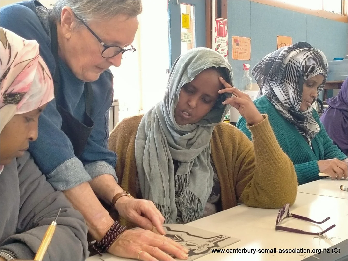 A diverse group of women sitting at a table, with an older woman helping a woman with a headscarf, while others observe and take notes.
