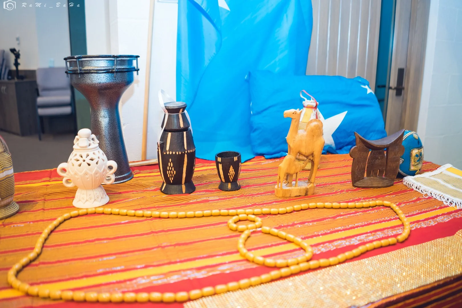 Collection of decorative items on a red textured cloth, including a tall black vase, a smaller black vase, a white ceramic urn, a carved wooden camel figurine, a wooden stool, and an orange beaded necklace, with a large blue cloth with star designs in the background.