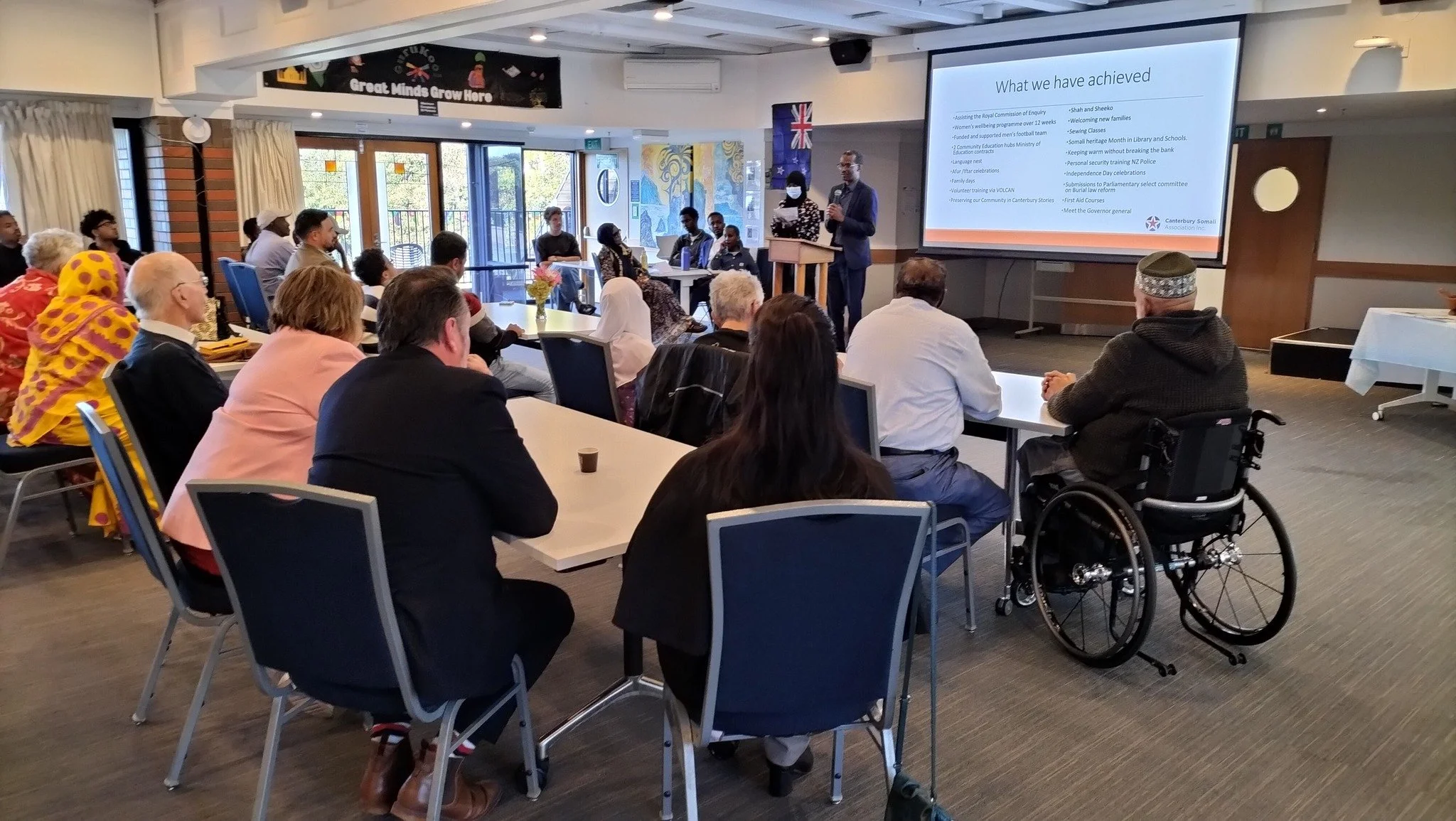 A diverse group of people attending a presentation in a conference room with a large screen displaying bullet points titled 'What we have achieved'. The speaker is standing at the front, and some attendees are wearing masks. There is a wheelchair person in the front right, and the room has large windows with natural light and decorative art on the walls.