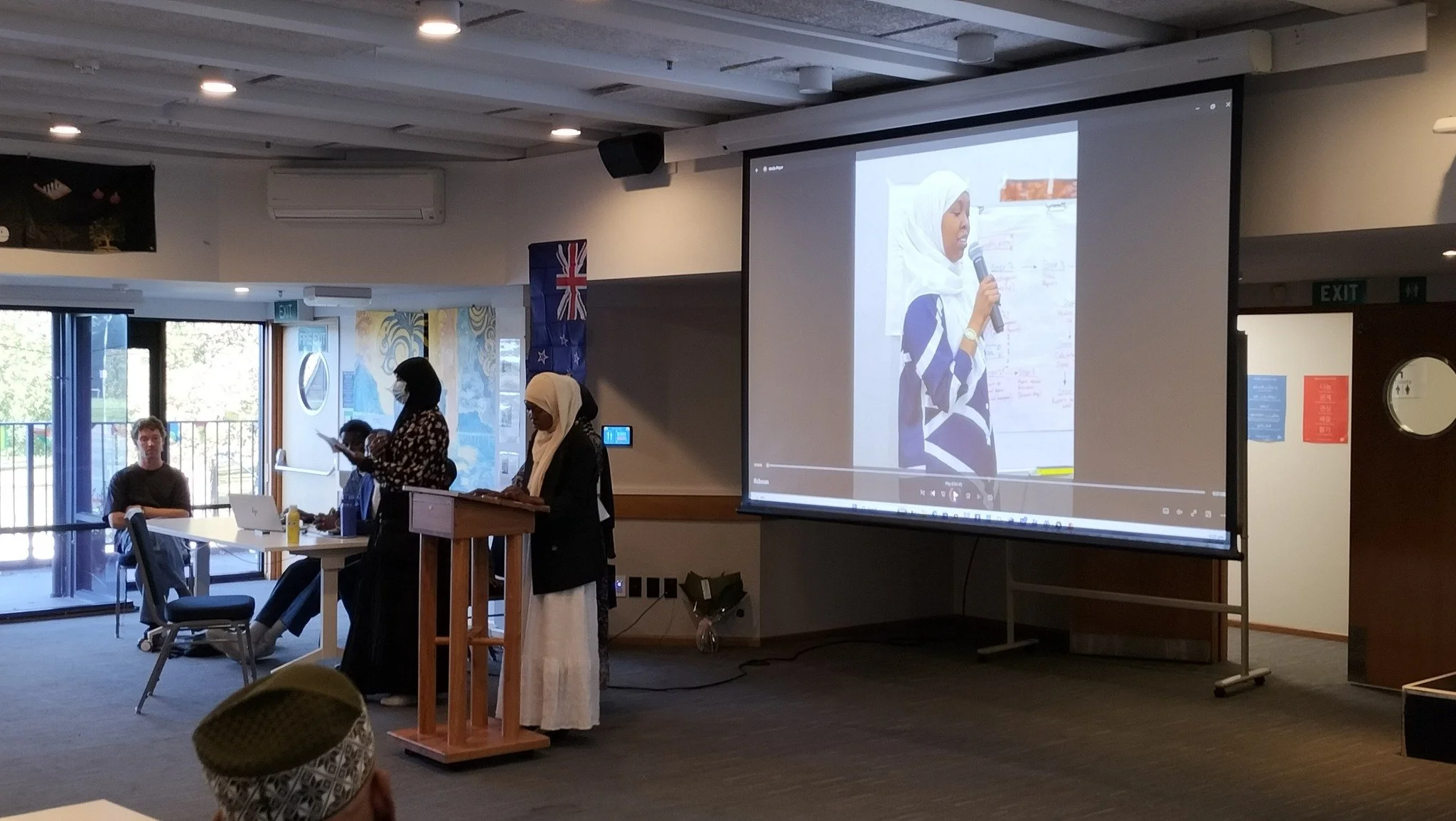Indoor classroom or meeting room with a presentation screen, two women of Middle Eastern or South Asian descent standing at a wooden podium, one speaking into a microphone, and a woman in a hijab sitting at a table.