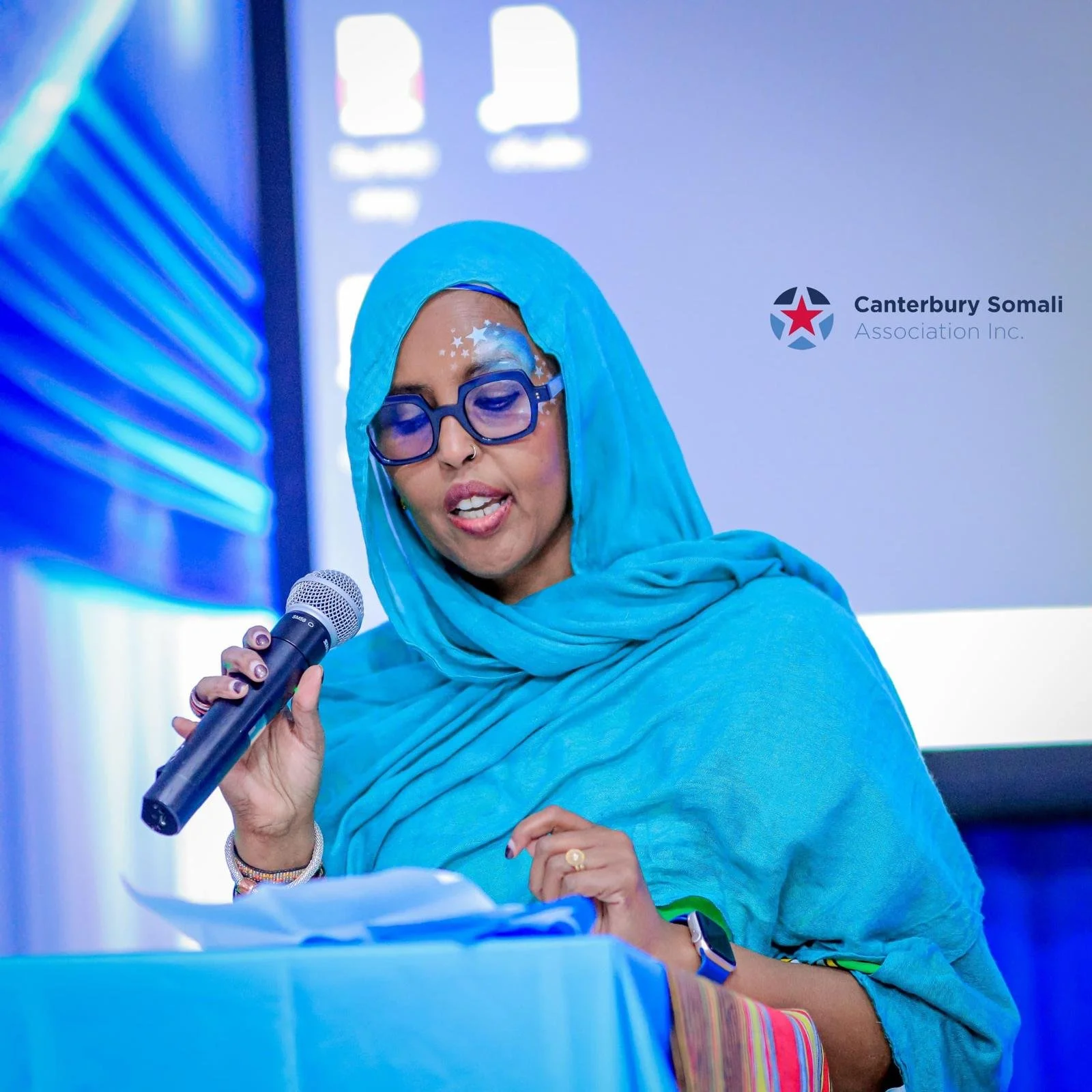 A woman wearing a blue hijab, glasses, and jewelry speaking into a microphone during a presentation or event. The background shows a logo for Canterbury Somali Association Inc.