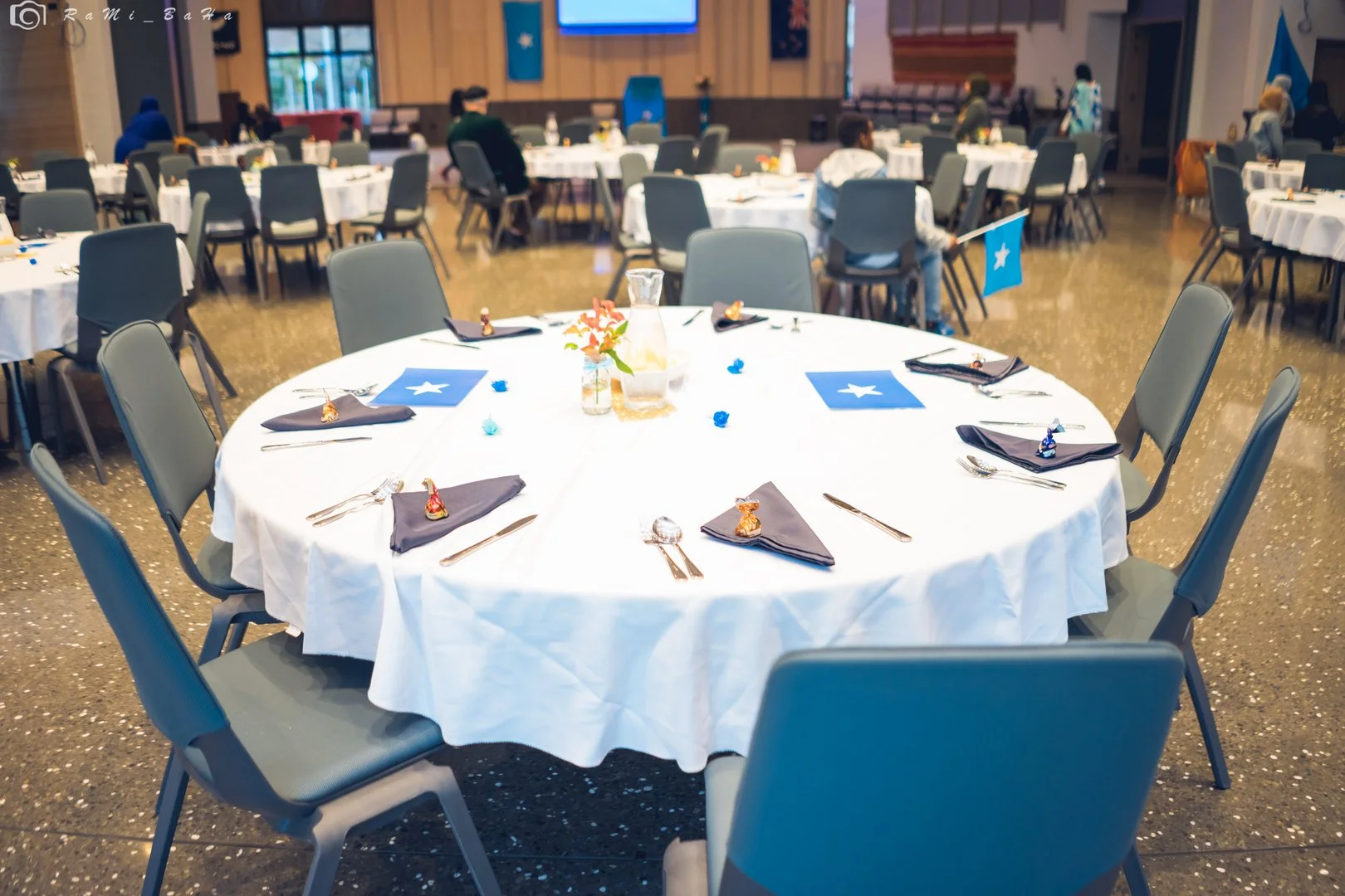 Round banquet table set with white tablecloth, blue napkins, silverware, and blue flags with a white star, in a decorated event hall with chairs around it and more tables in the background.