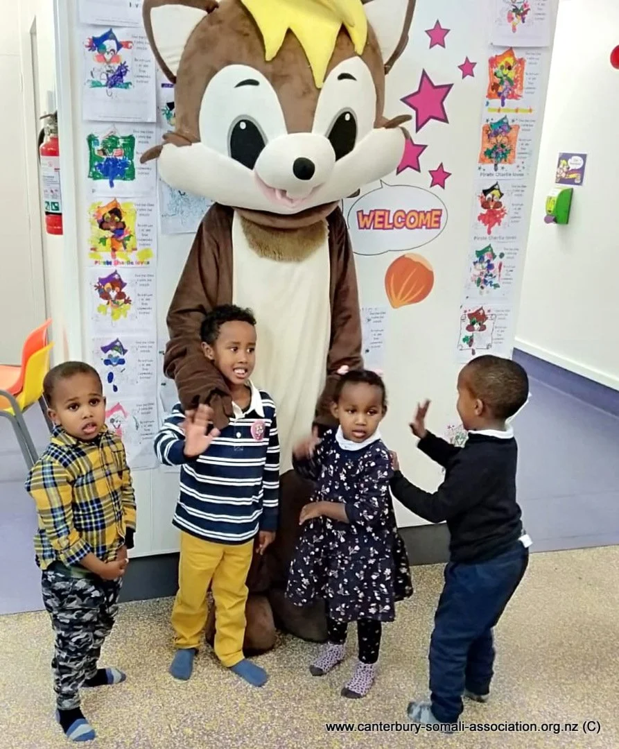 Four children standing in front of a person dressed in a large chipmunk costume. The children appear to be talking or interacting with the character, which is in front of a decorated wall with colorful drawings and a 'Welcome' sign.