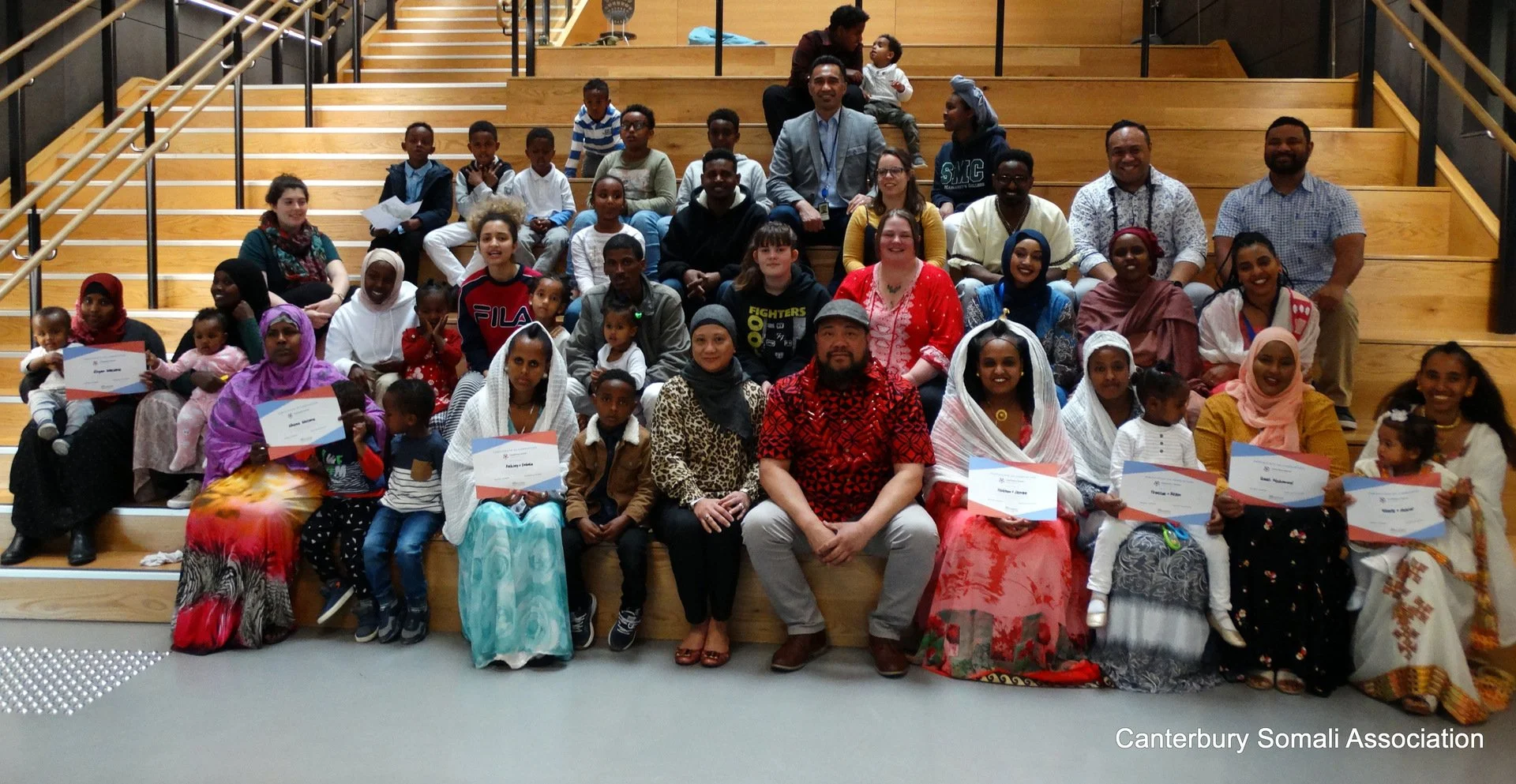 Group photo of diverse adults and children sitting on wooden stairs inside a building, some holding certificates, at an event organized by Canterbury Somali Association.