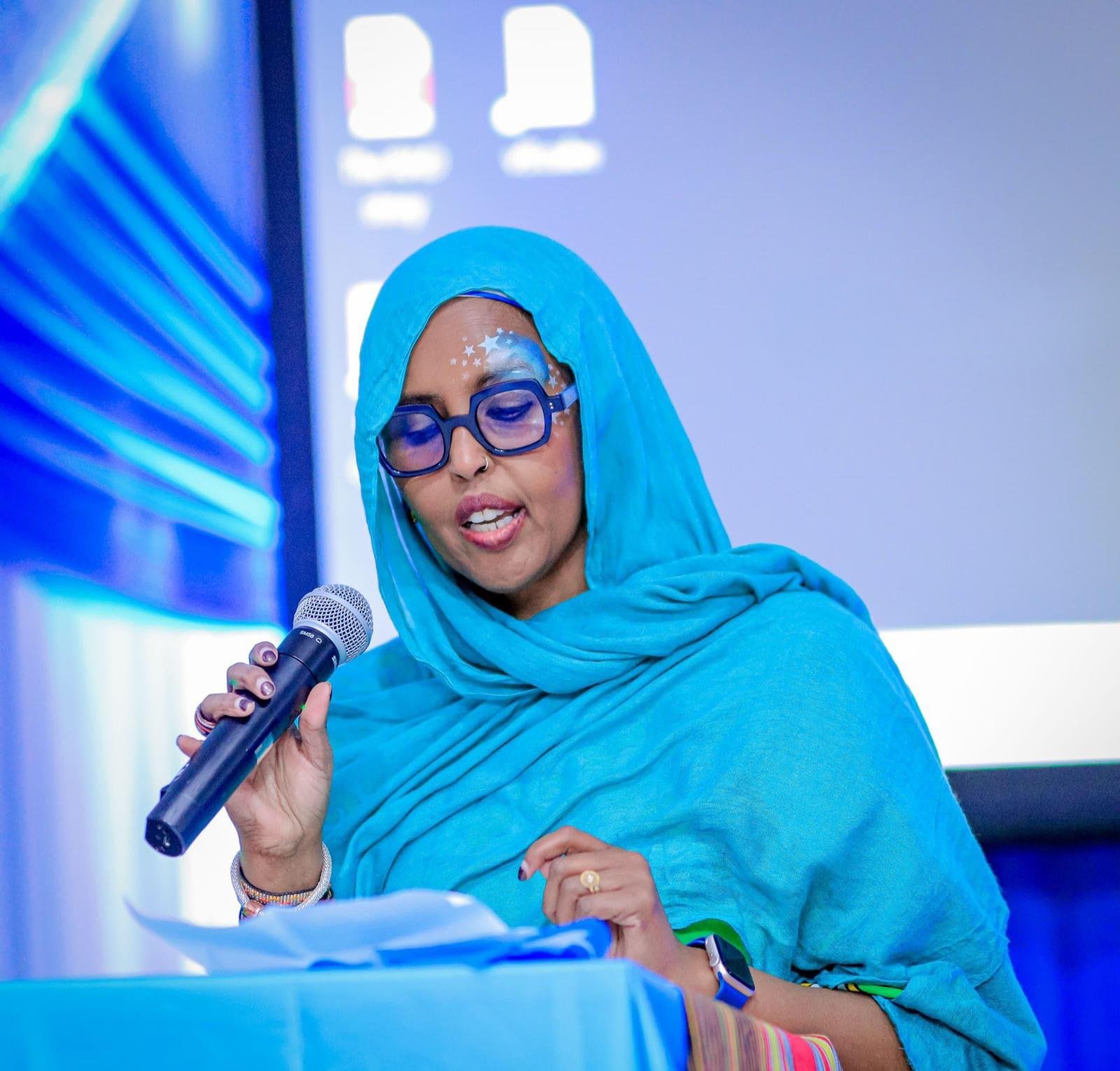 A woman wearing a blue hijab, glasses, and jewelry speaking into a microphone during a presentation or event. The background shows a logo for Canterbury Somali Association Inc.