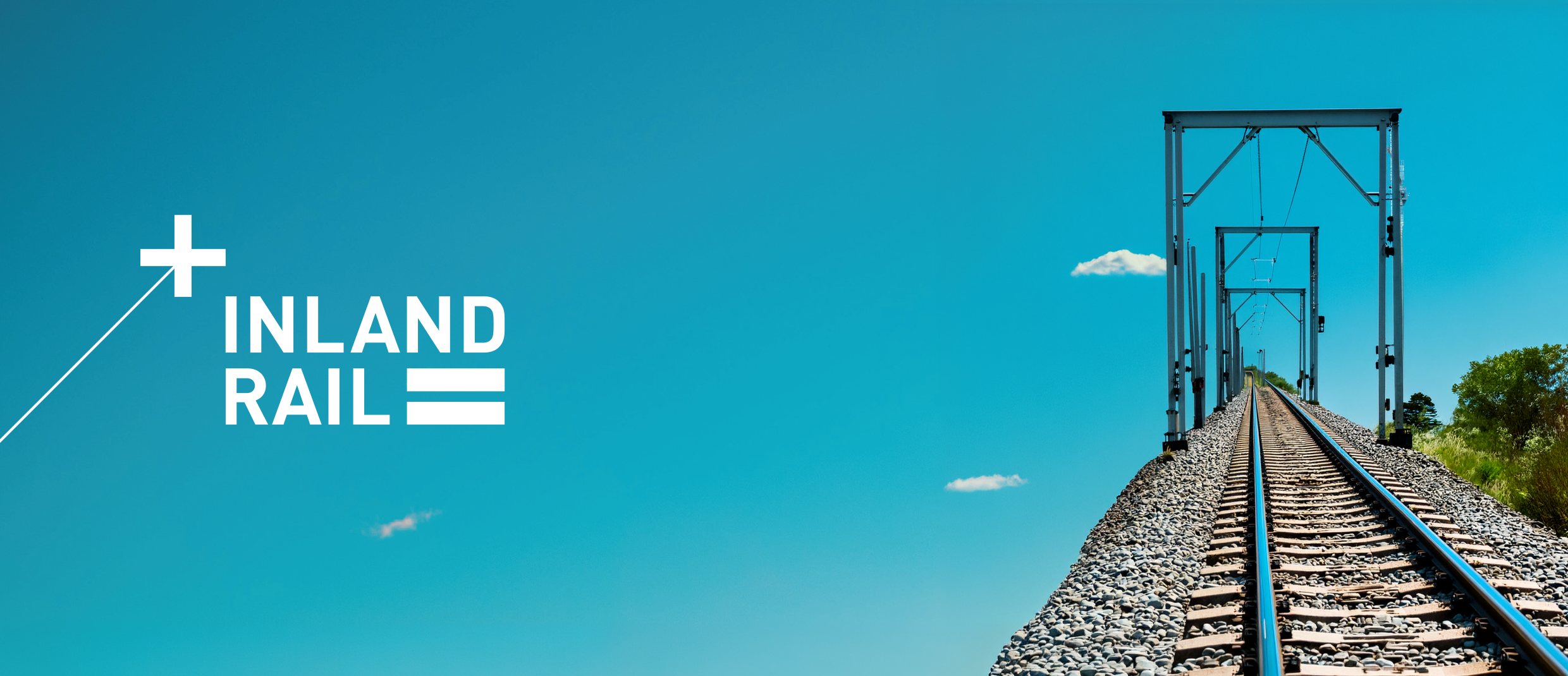 A railway track with overhead structures extending into the distance under a clear blue sky with a few clouds and some green trees on the right side.