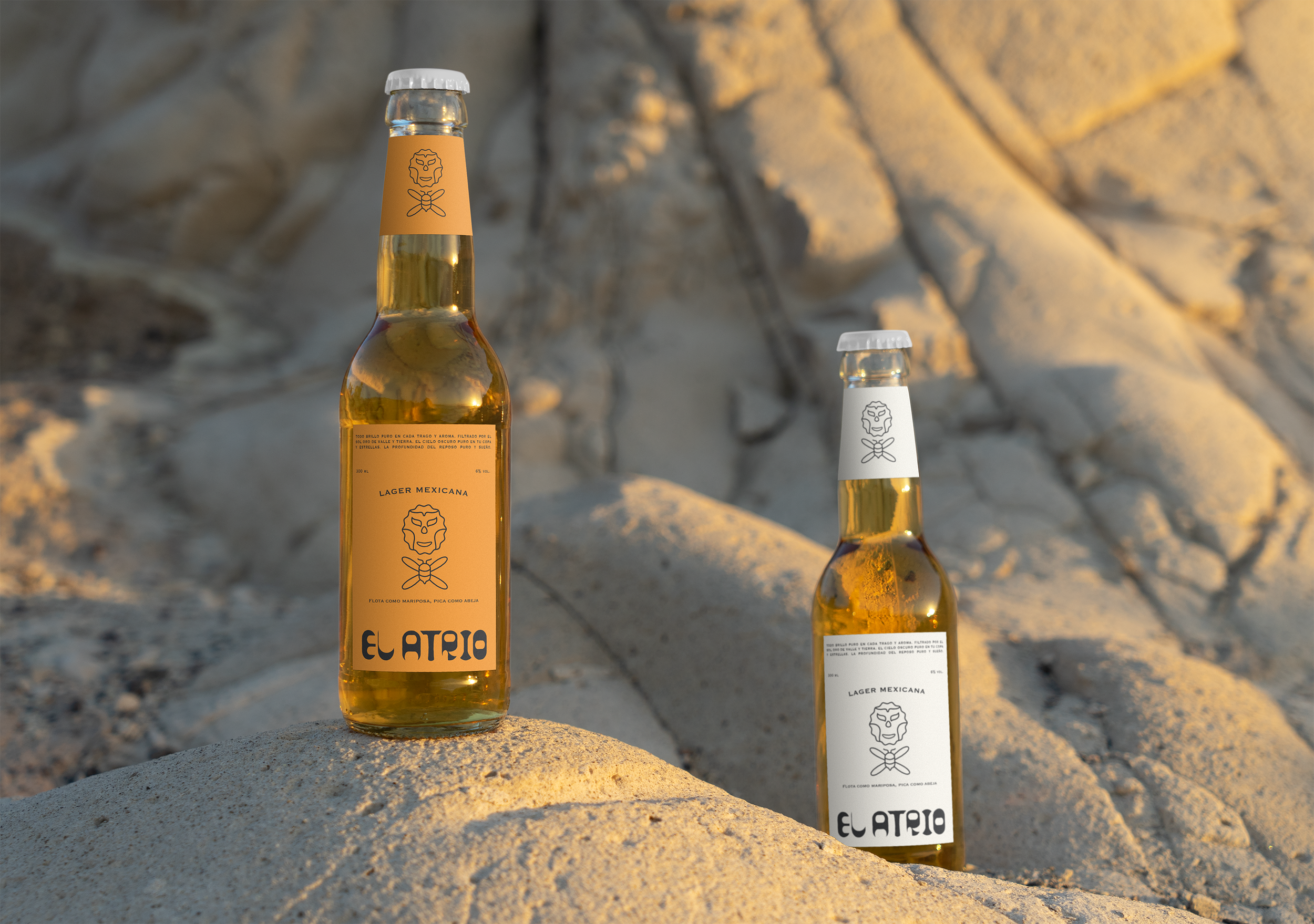 Two bottles of El Altrio Mexican lager beer placed on sandy rocks, one with a beige label and the other with a white label, in an outdoor setting with rocky terrain in the background.