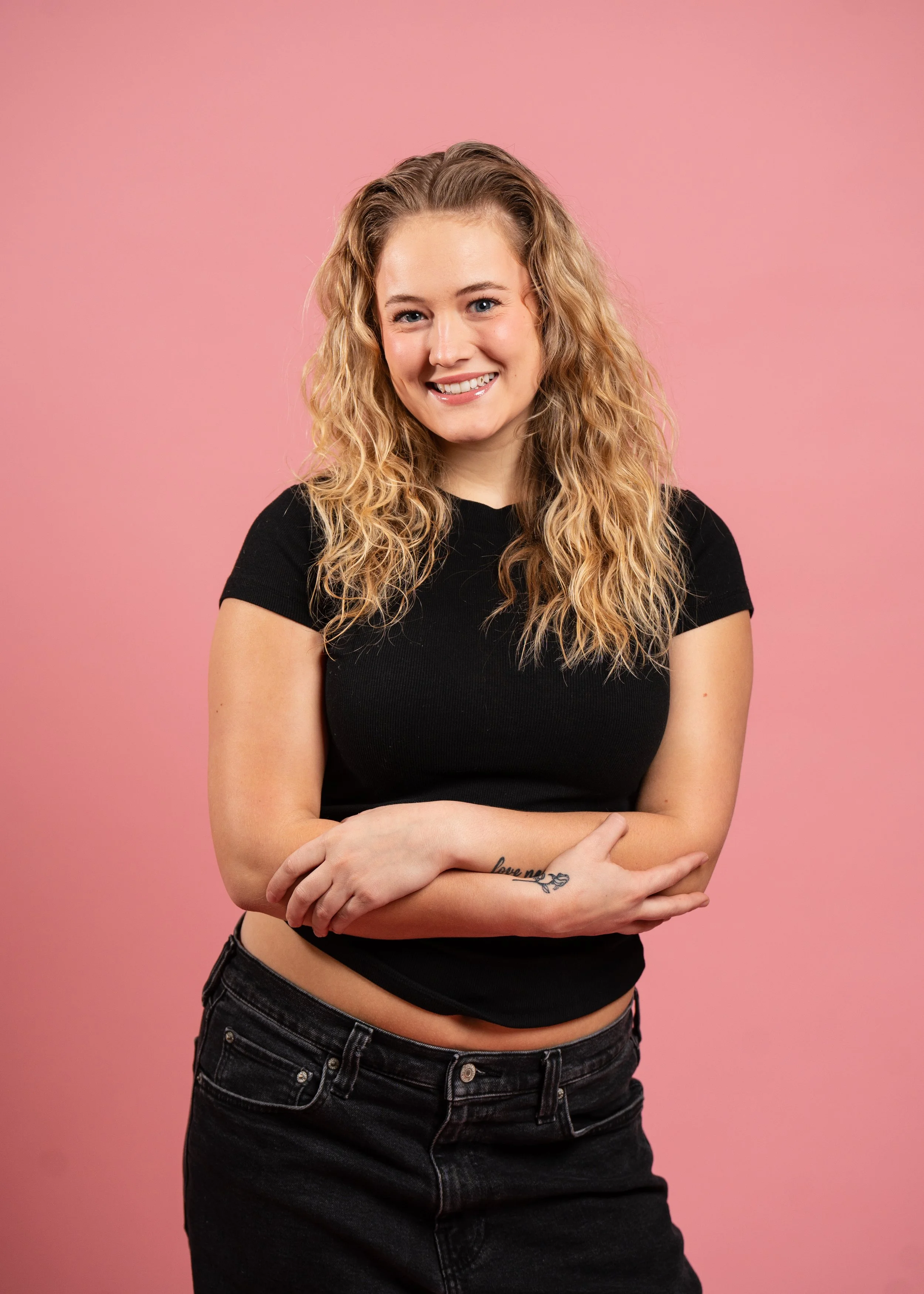 A young woman with curly blonde hair smiling, wearing a black t-shirt and black jeans, standing against a pink background. She has crossed arms with a tattoo that says 'love me' on her forearm.
