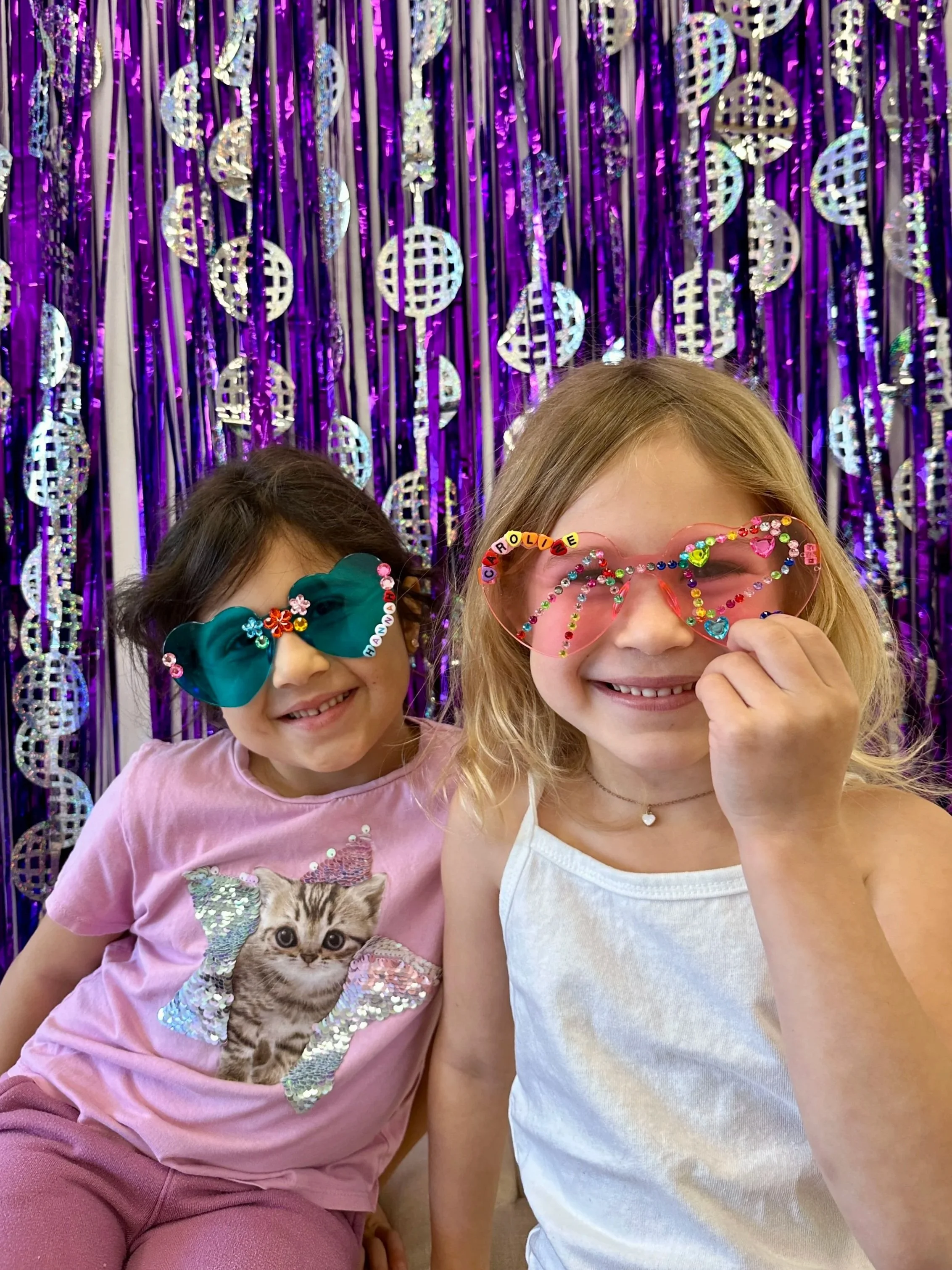 Two young girls smiling and wearing decorated glasses in front of a purple tinsel curtain with disco ball decorations.