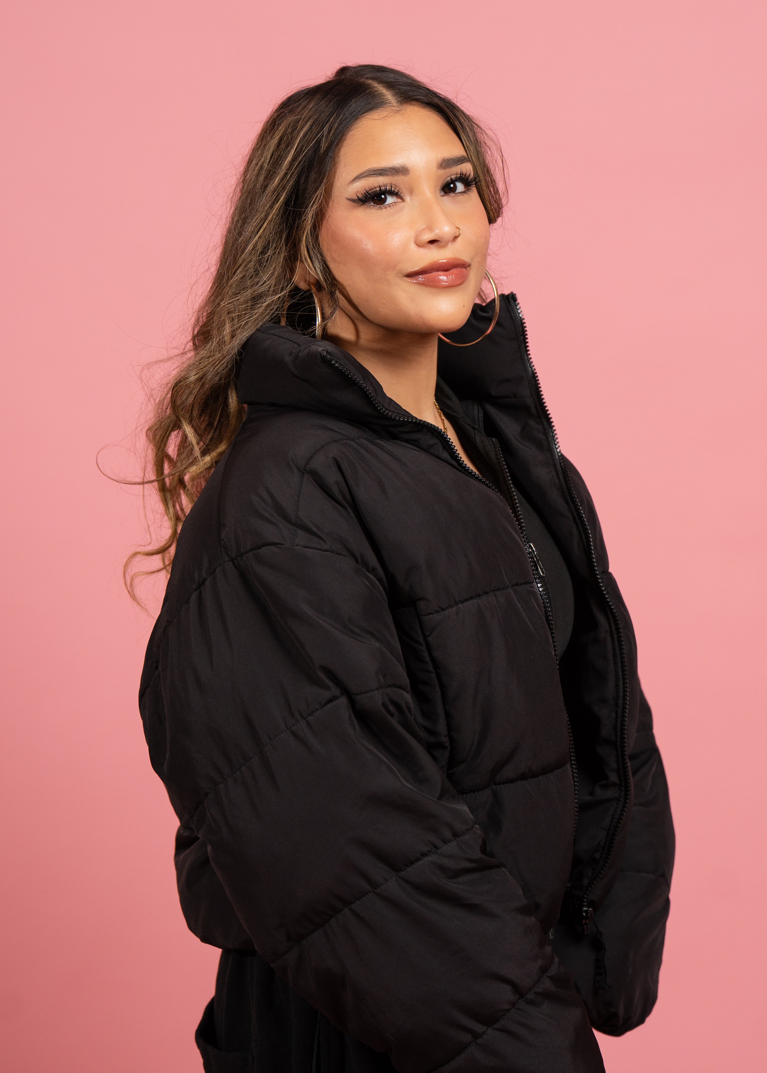 A young woman with long wavy hair, wearing large hoop earrings, glossy makeup, and a black puffer jacket, standing against a pink background.