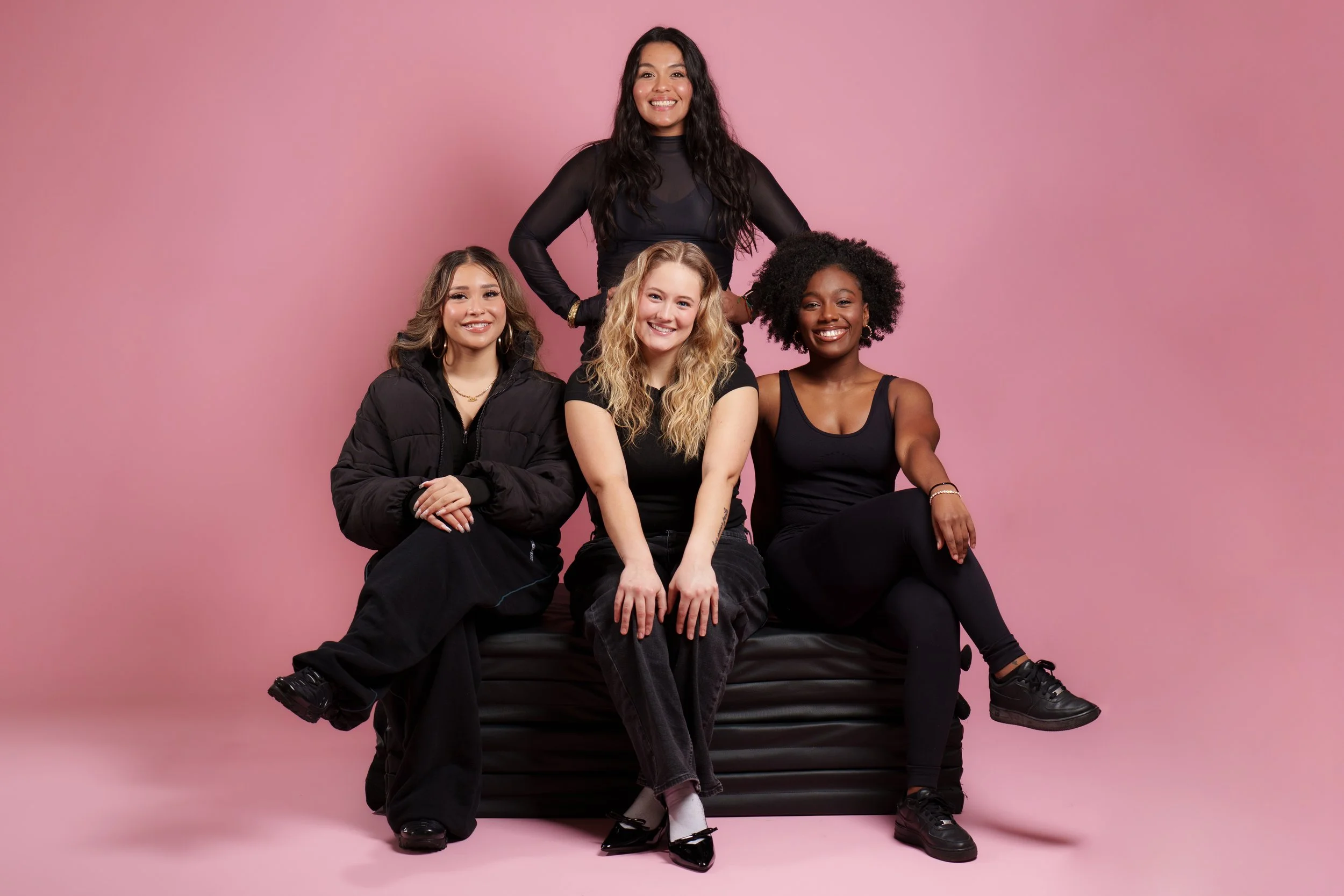 Five smiling women sitting and standing on a black leather sofa against a pink background, dressed casually in black clothes.
