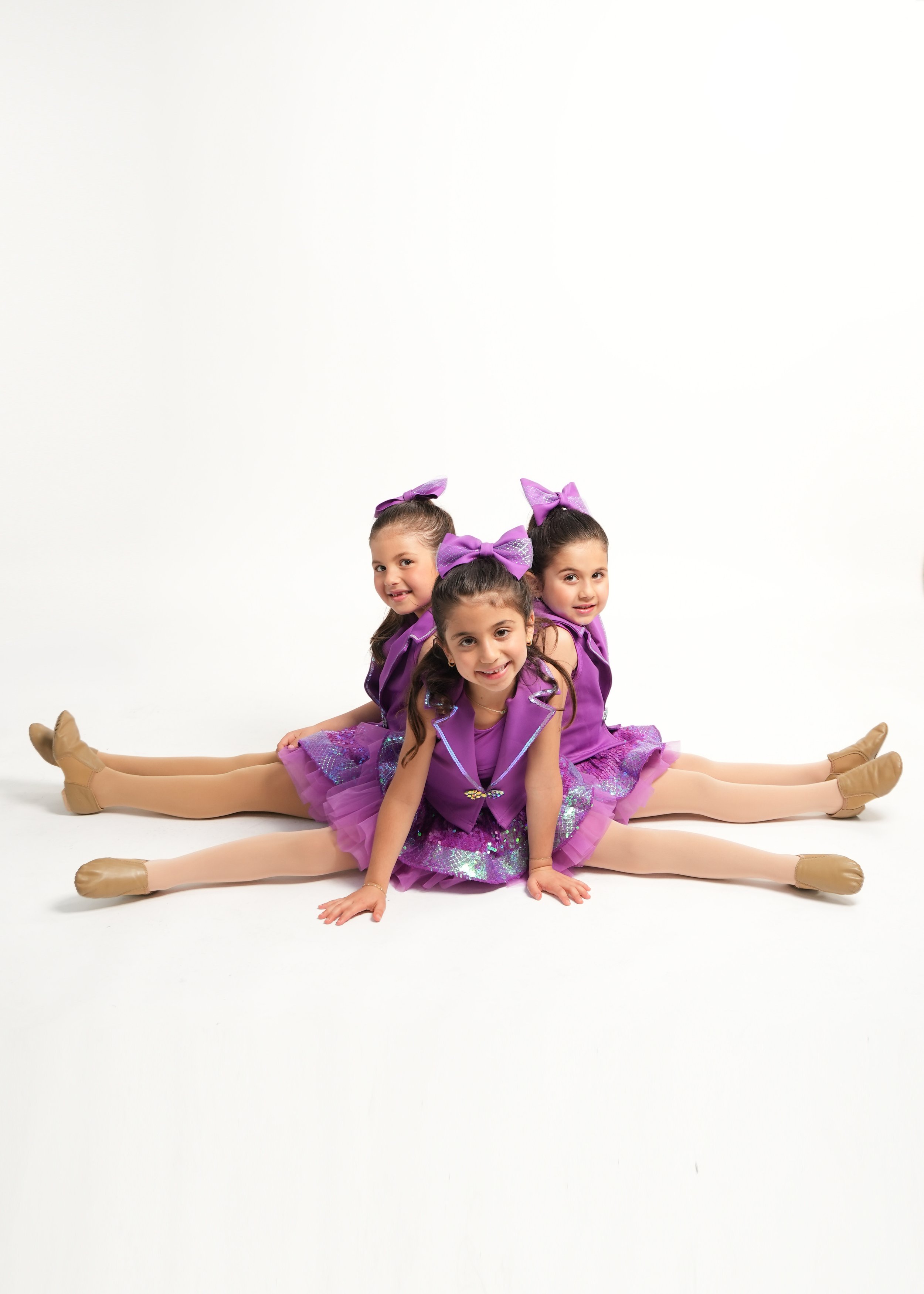 Three young girls in purple dance costumes and ballet shoes performing a split on a white background.