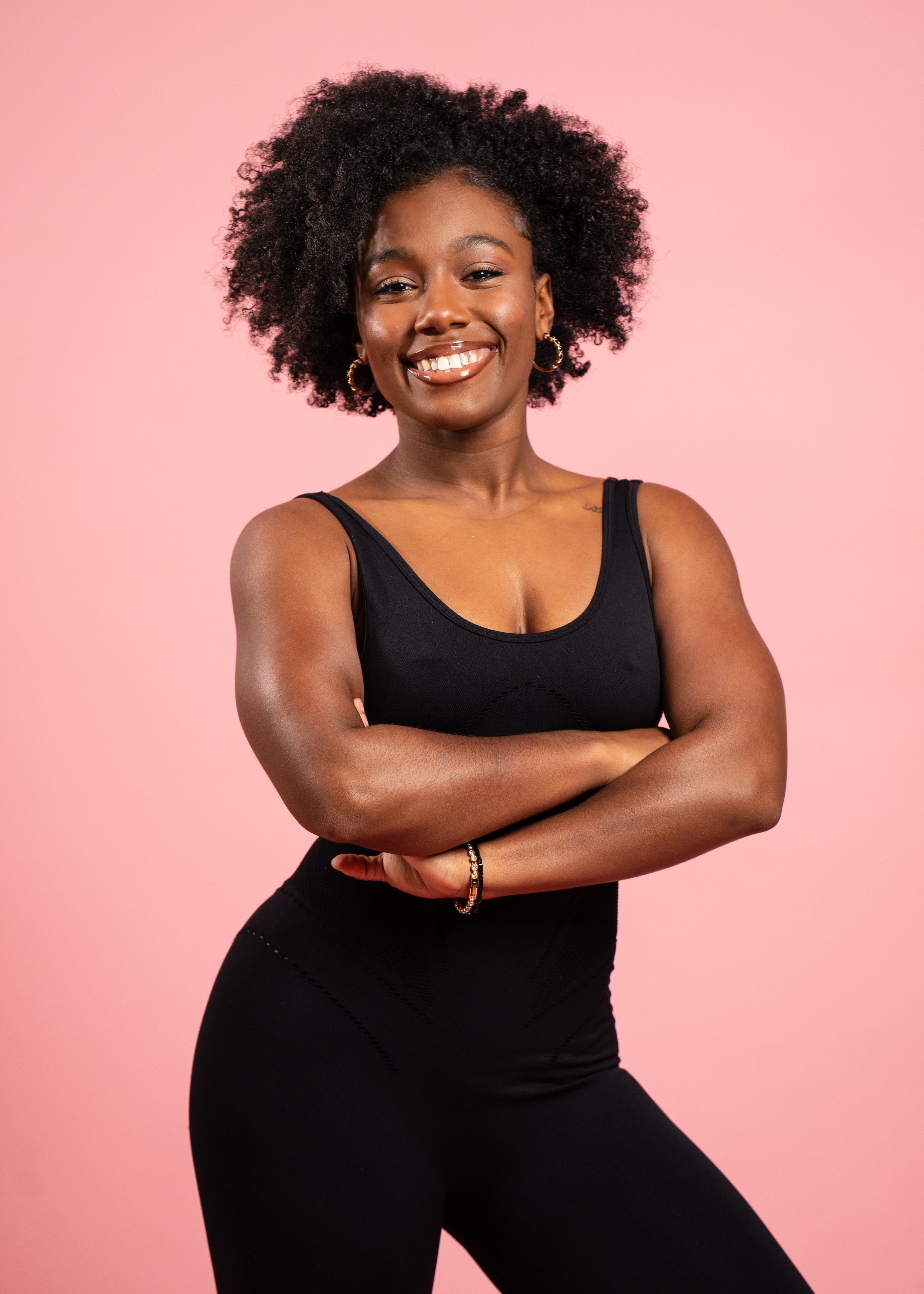 A smiling Black woman with curly hair, wearing a black athletic tank top and black leggings, standing with arms crossed in front of a pink background.