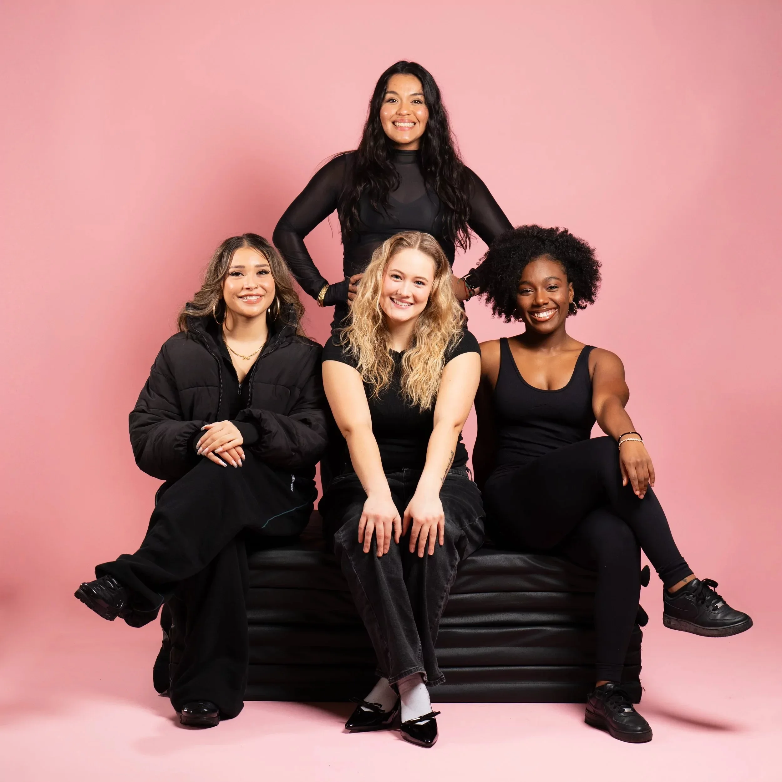Group of five diverse women posing together against a pink background, sitting and standing on a black sofa, smiling.
