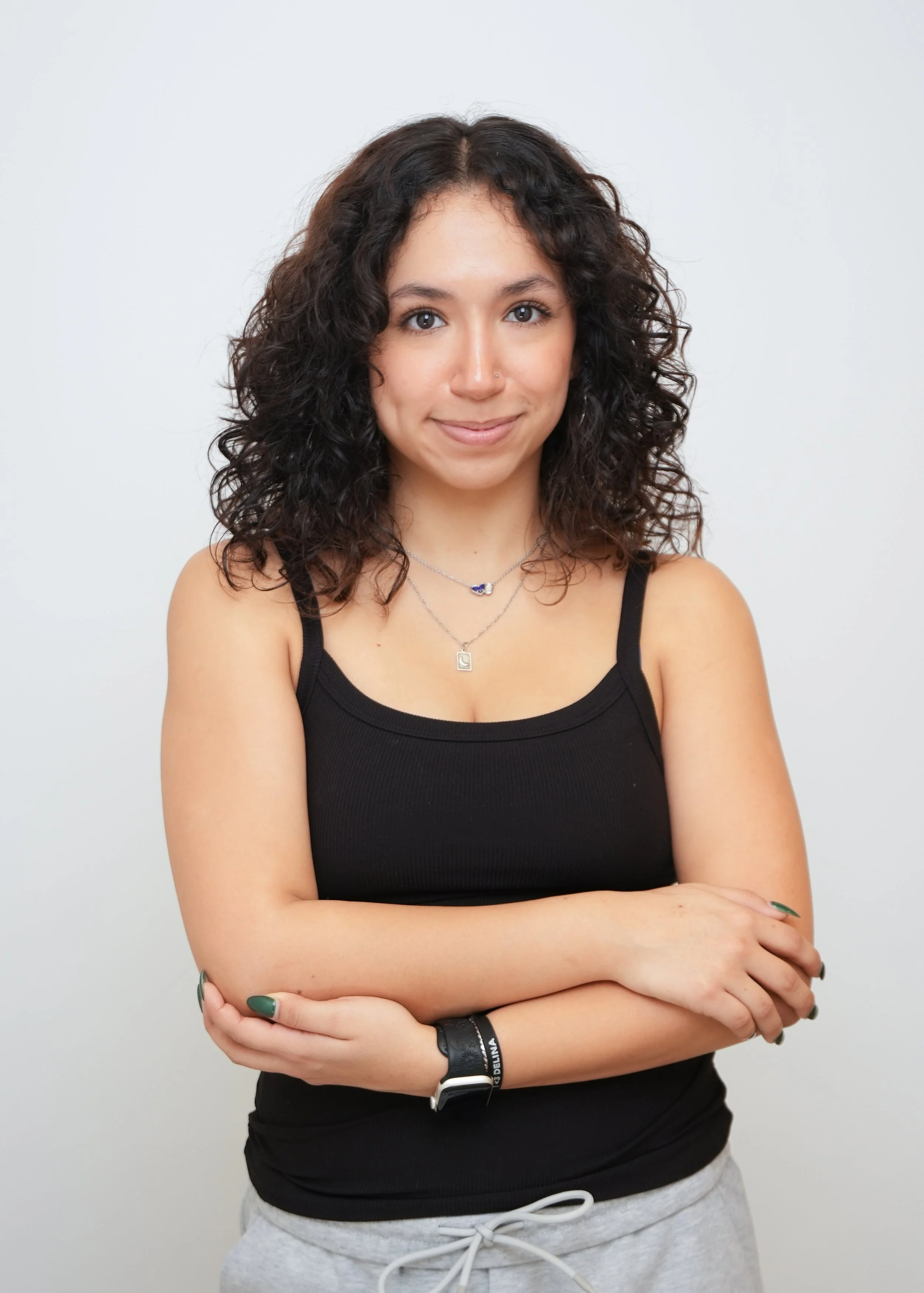 A young woman with curly dark hair wearing a black tank top and gray sweatpants, standing against a white background with her arms crossed, smiling softly at the camera.