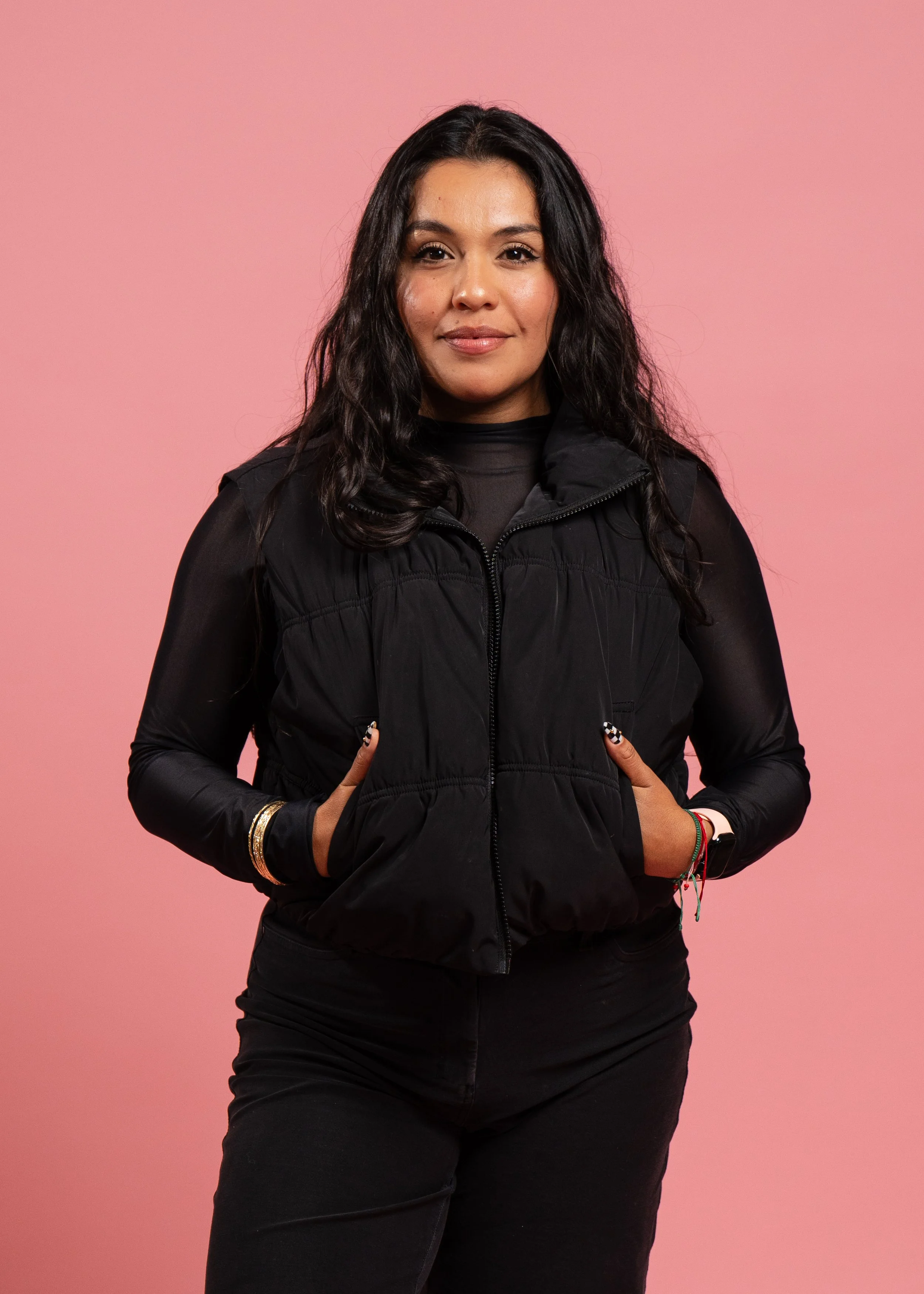 A woman with long black wavy hair, wearing a black vest over a long sleeve black shirt, standing against a pink background with her hands in her pockets.