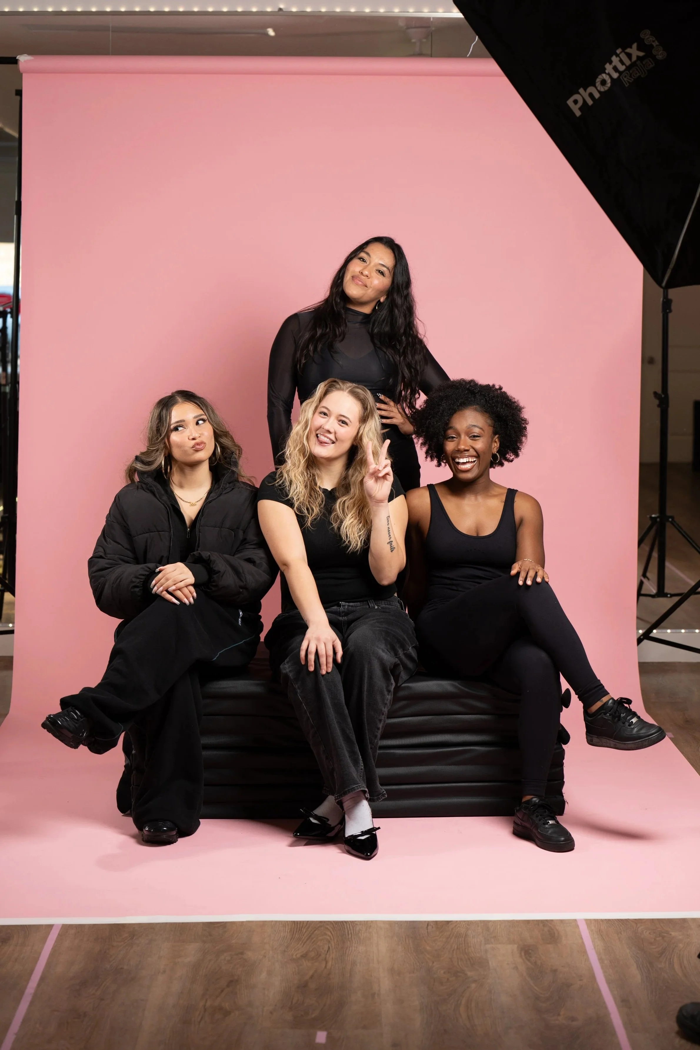 Group of four women posing together in front of a pink backdrop at a photo shoot, with two sitting on a black bench and two standing behind. They are all smiling and dressed in black or dark clothing.