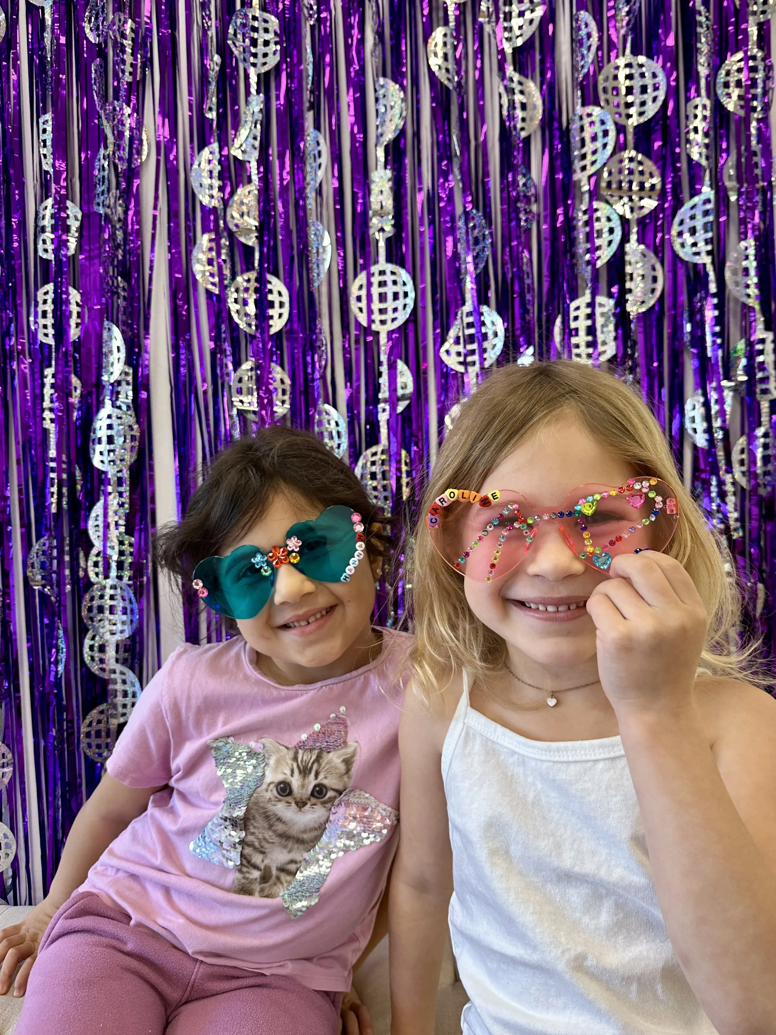 Two young girls smiling, wearing colorful sunglasses with decorated frames, standing in front of a purple metallic streamers backdrop. One girl has long blonde hair and is wearing a white top, the other girl has short dark hair and is wearing a pink shirt with a cat graphic.