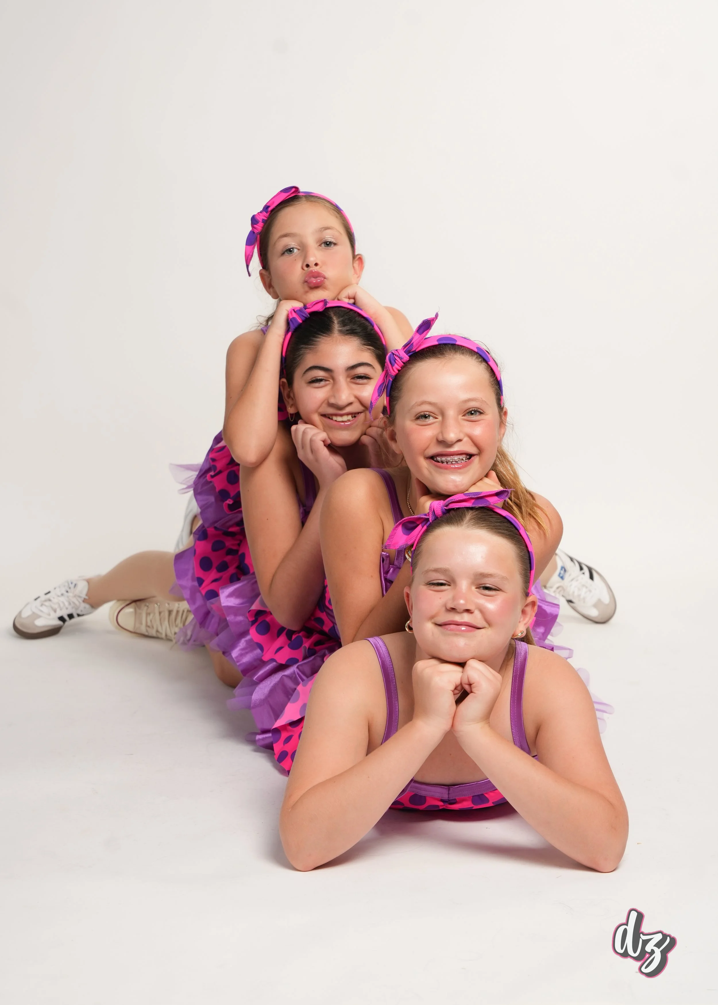 Four young girls in colorful dance costumes and matching headbands lying on the floor in a line, smiling and posing for the camera against a plain white background.