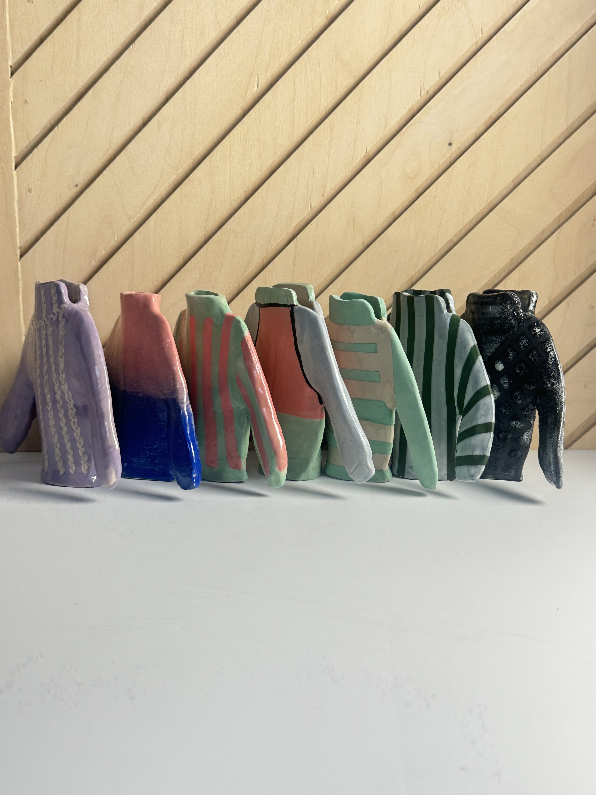 Colorful ceramic sculptural sweater bud vases, arranged in a row on a white surface against a wooden board background.