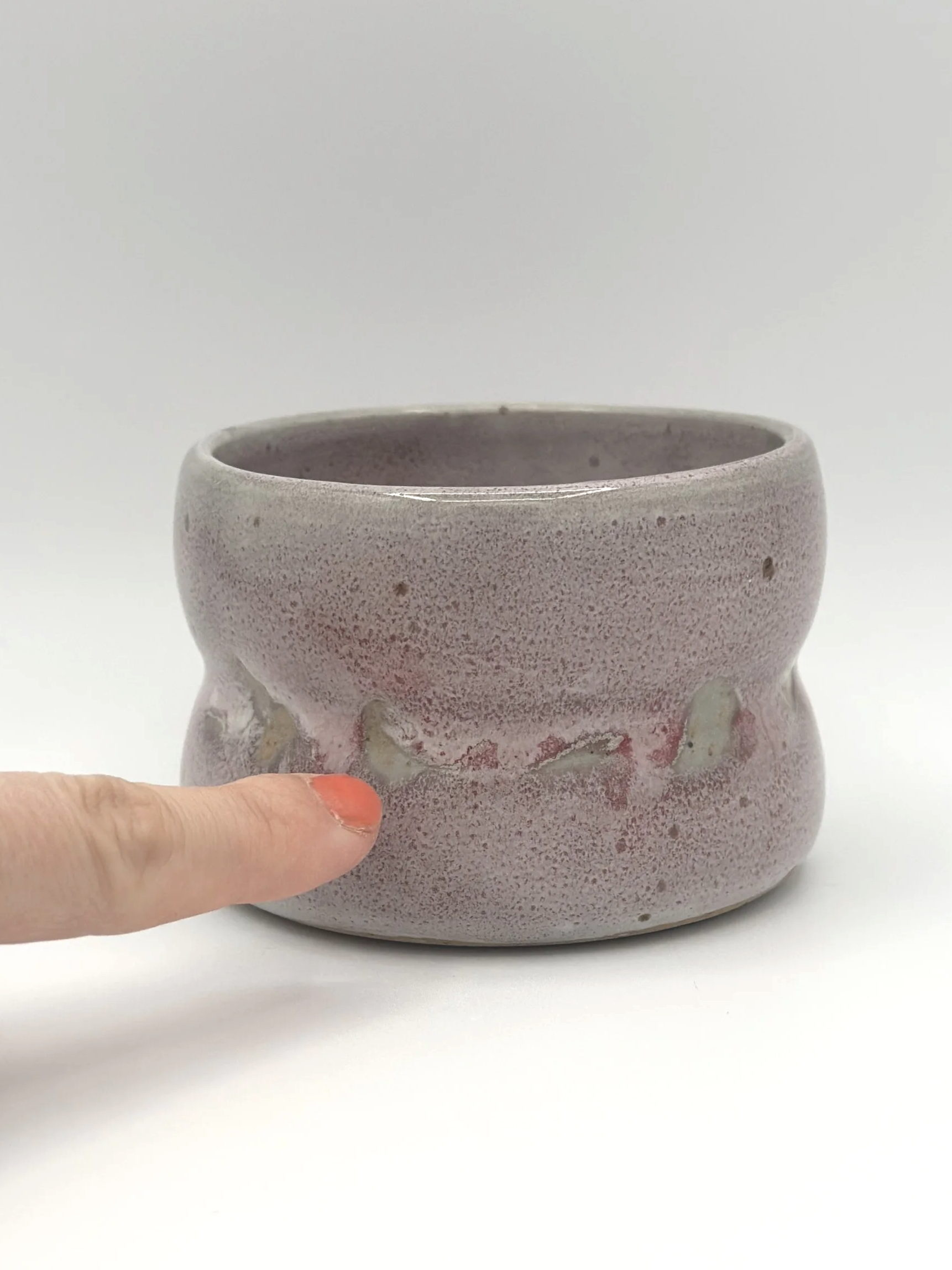 Handmade stoneware ceramic tumbler with 2 bubbles and a light grayish and pinkish candy coated shiny glaze. manicured finger pointing to glaze imperfections