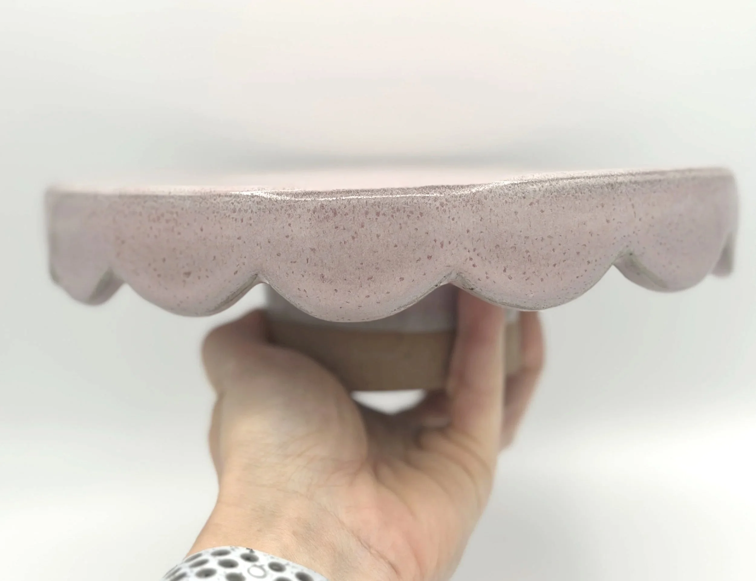 handmade stoneware ceramic cake stand with scalloped edges and pink speckled glaze held by the bottom in a hand