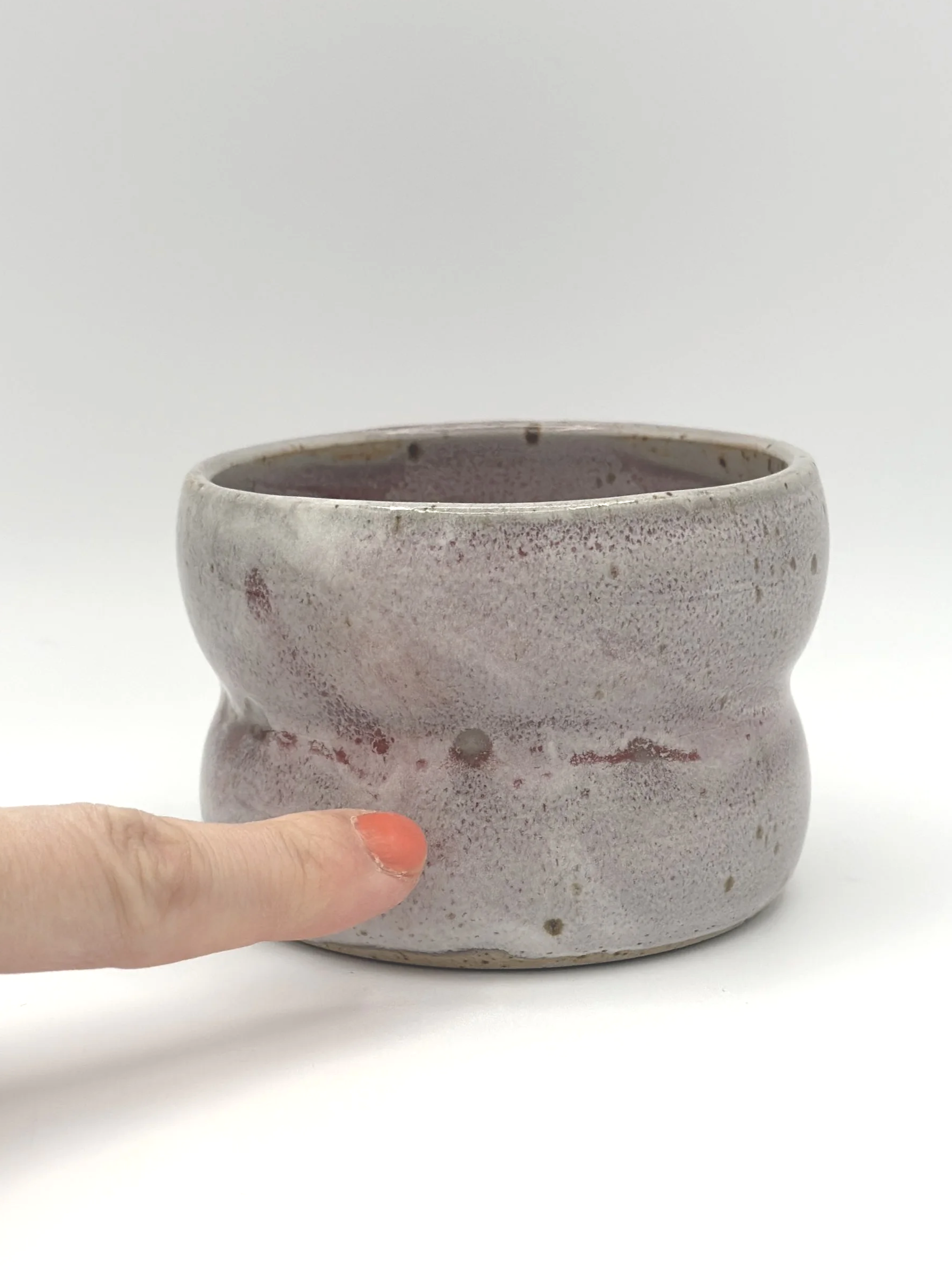 Handmade stoneware ceramic tumbler with 2 bubbles and a light grayish and pinkish candy coated shiny glaze. manicured finger pointing to glaze imperfections