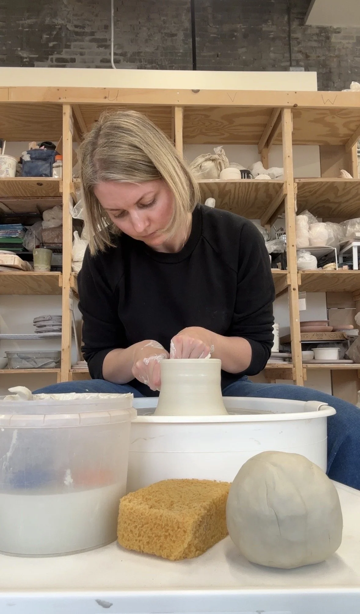 A woman working on pottery at a wheel in a ceramics studio. In the foreground, there is a sponge, a partially finished spherical clay form, and a container of water.