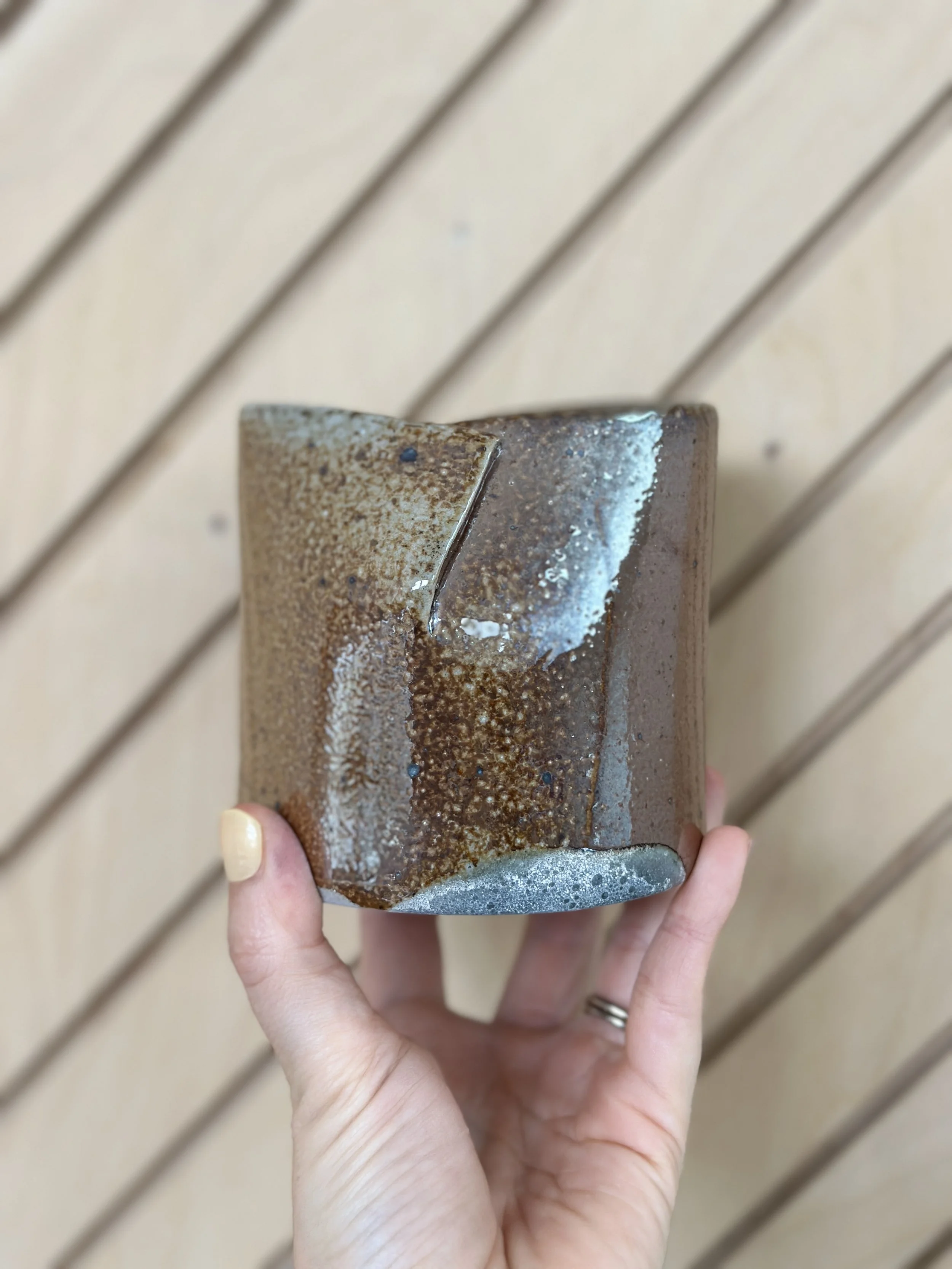 folded shiny metallic ceramic vessel held by a hand with a diagonal modern wood background