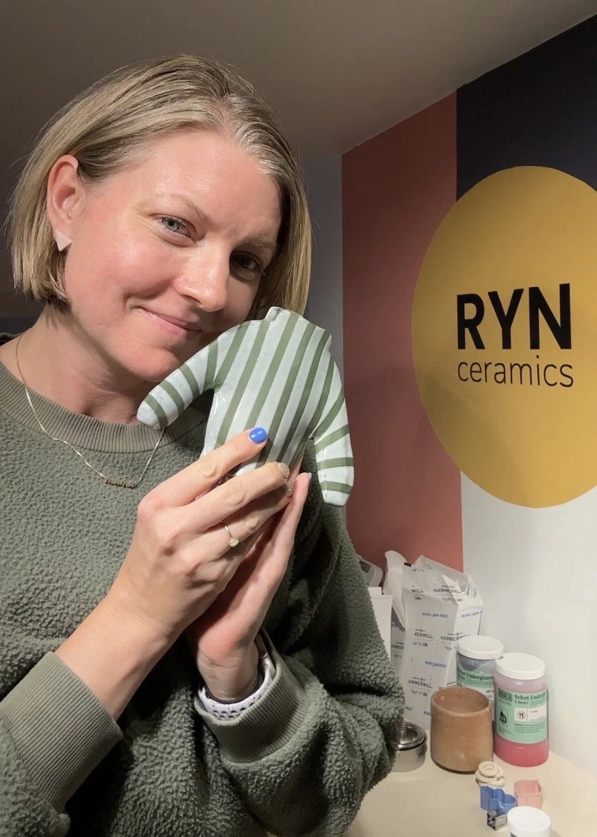 A woman with short blonde hair holding a ceramic piece, smiling at the camera, standing in front of a RYN ceramics sign.
