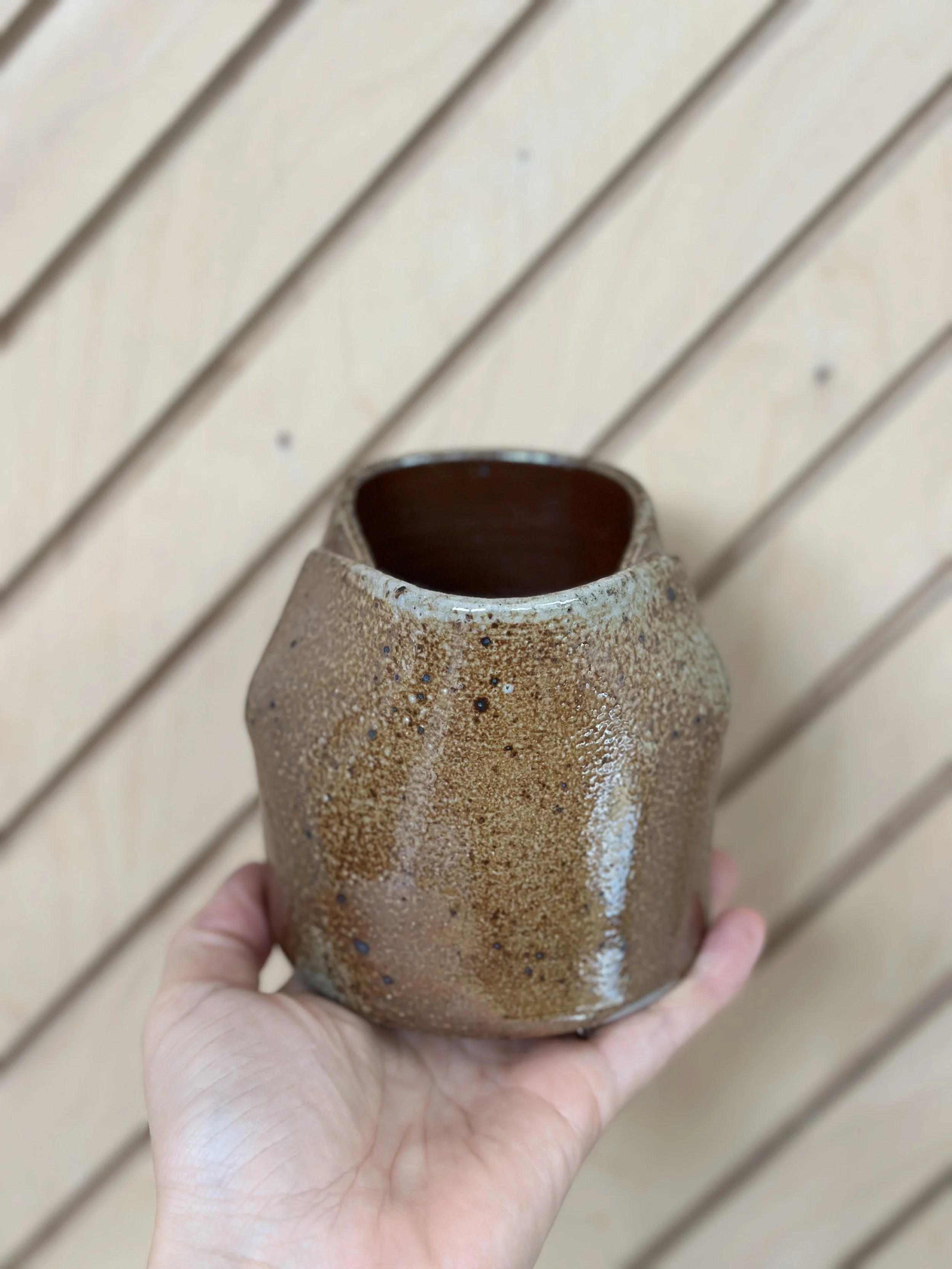 folded shiny metallic ceramic vessel held by a hand with a diagonal modern wood background