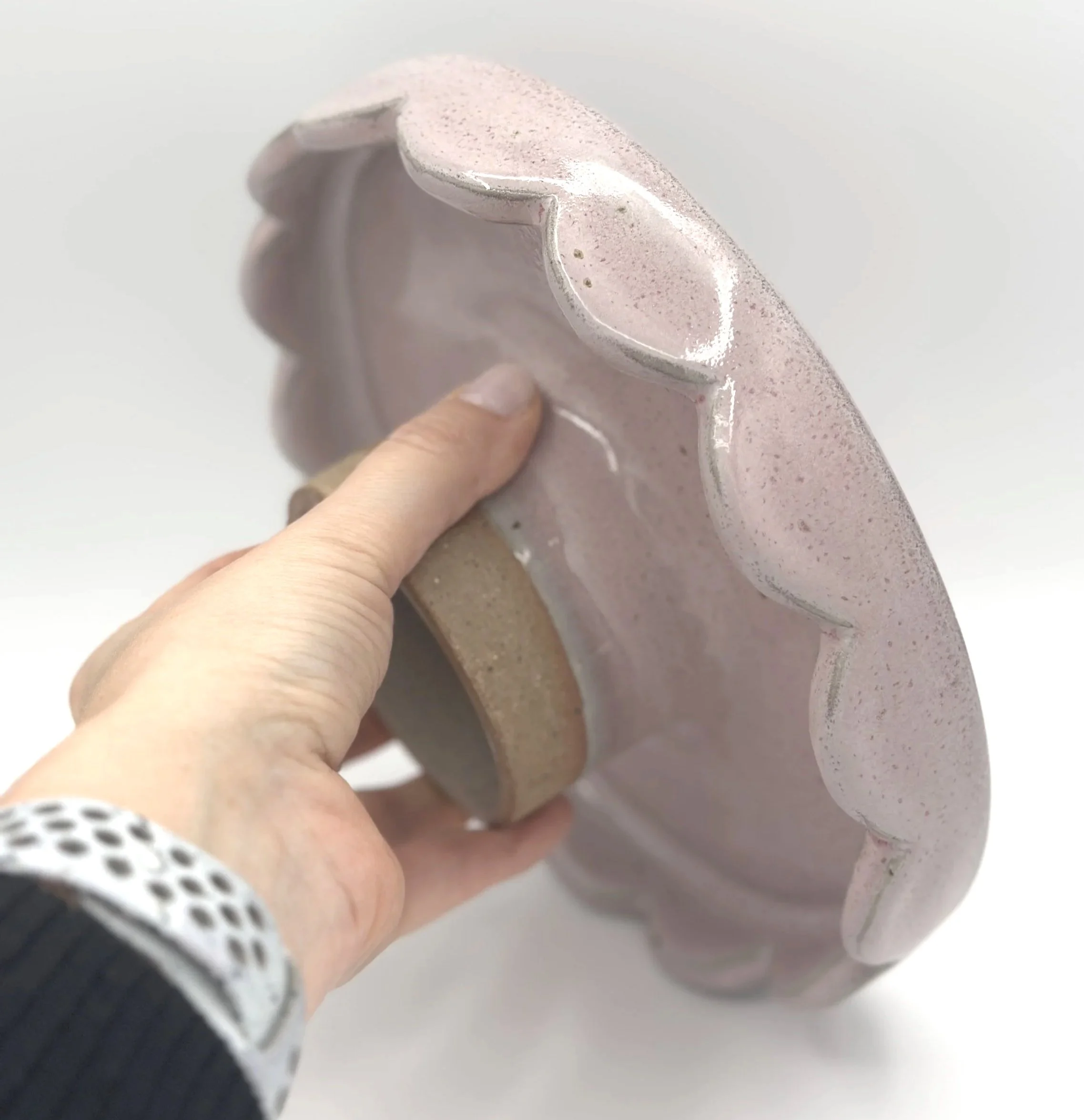 handmade stoneware ceramic cake stand with scalloped edges and pink speckled glaze held by the bottom in a hand showing off the small unglazed stem