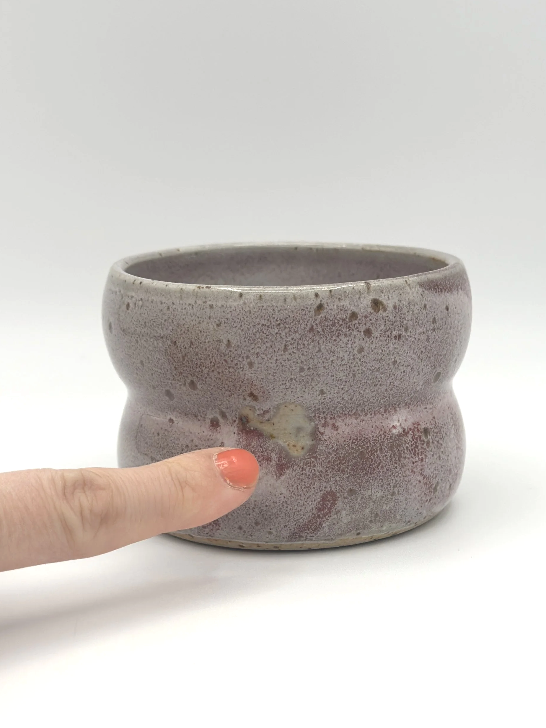 Handmade stoneware ceramic tumbler with 2 bubbles and a light grayish and pinkish candy coated shiny glaze. manicured finger pointing to glaze imperfections